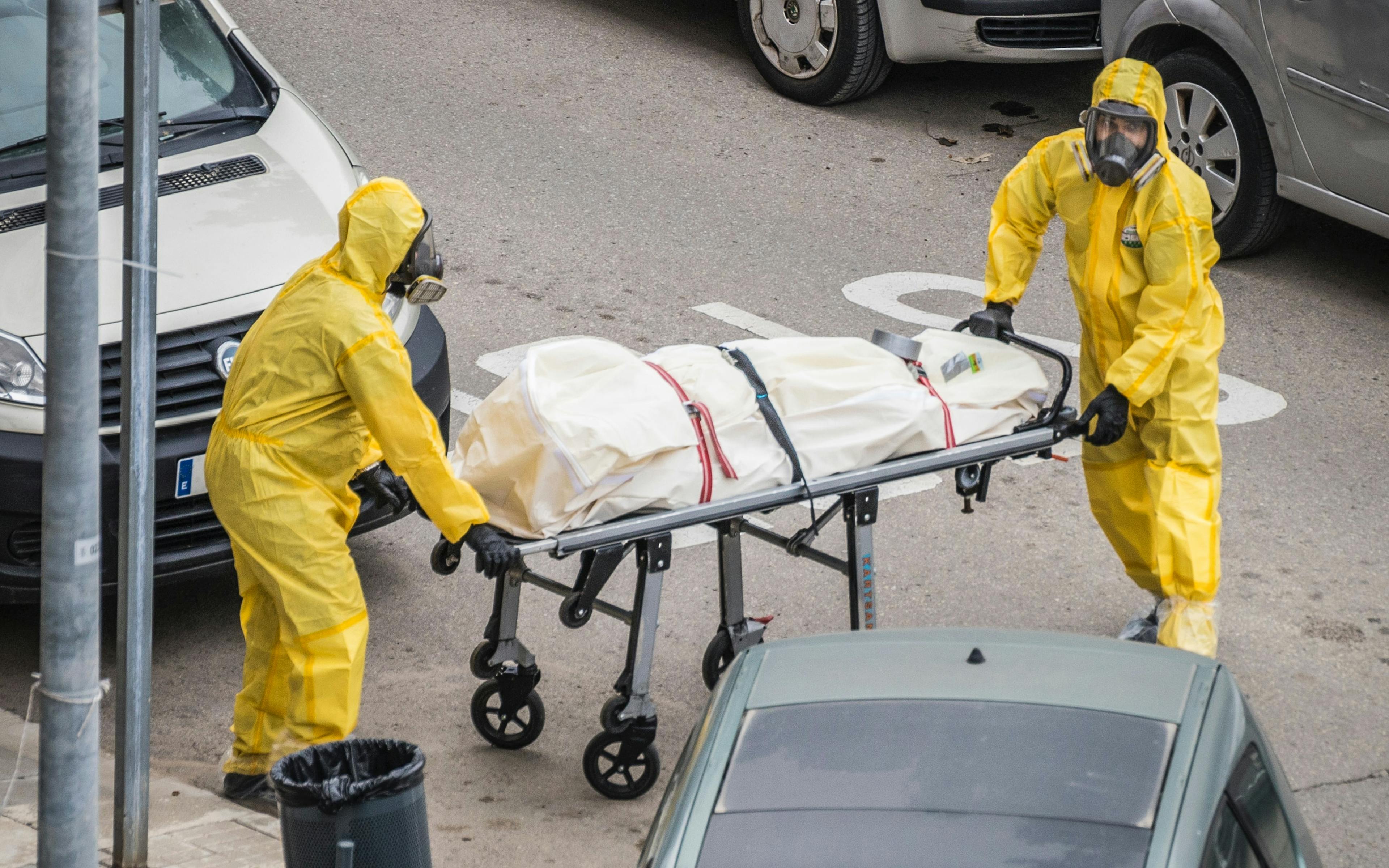 Two people in yellow PPE wheeling wrapped body through street
