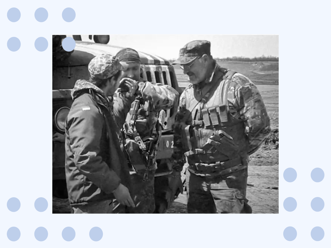 The image shows three soldiers talking in front of a military vehicle