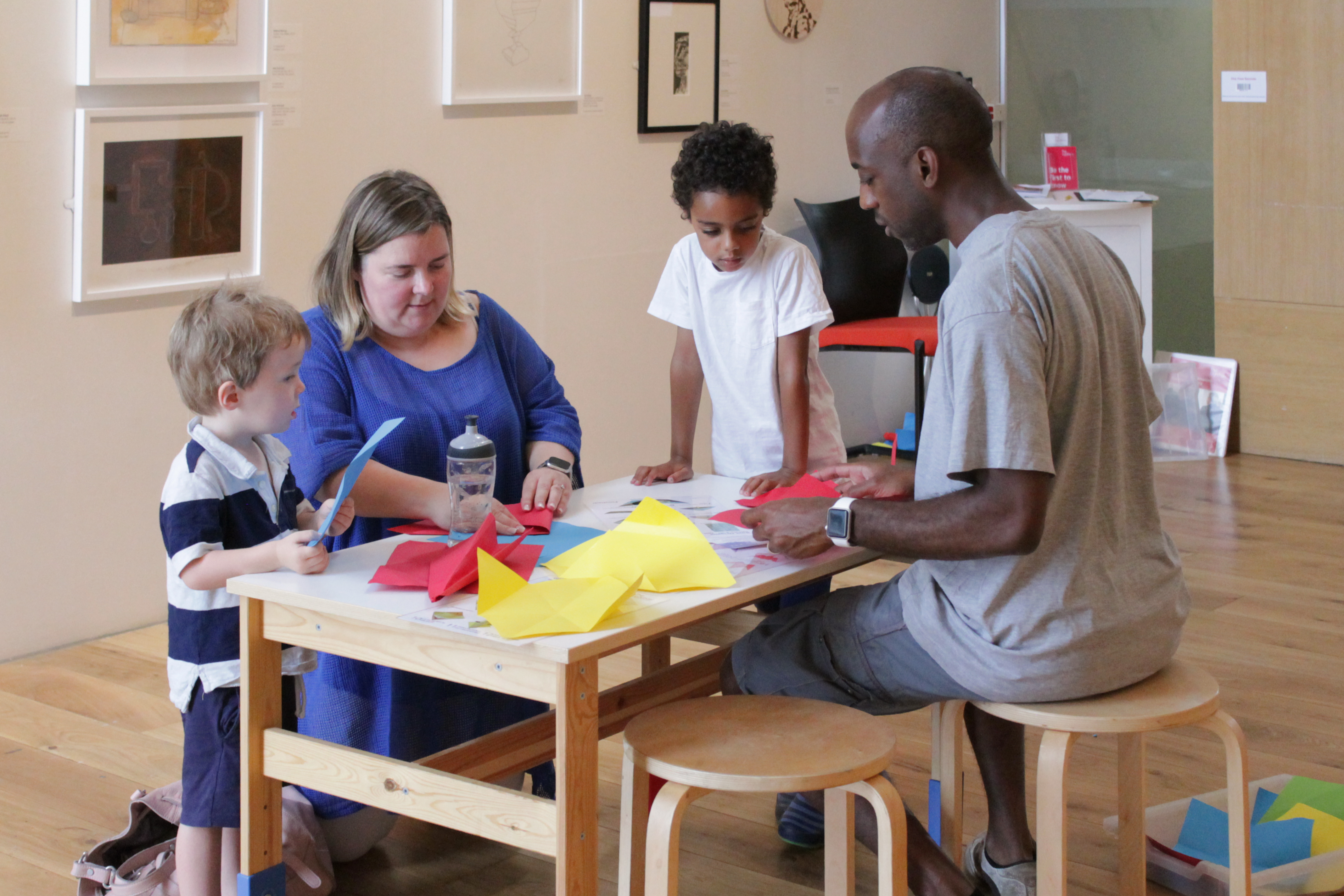 A family taking part in an arts and crafts work shop. Making paper airplanes out of colourful pieces of card.
