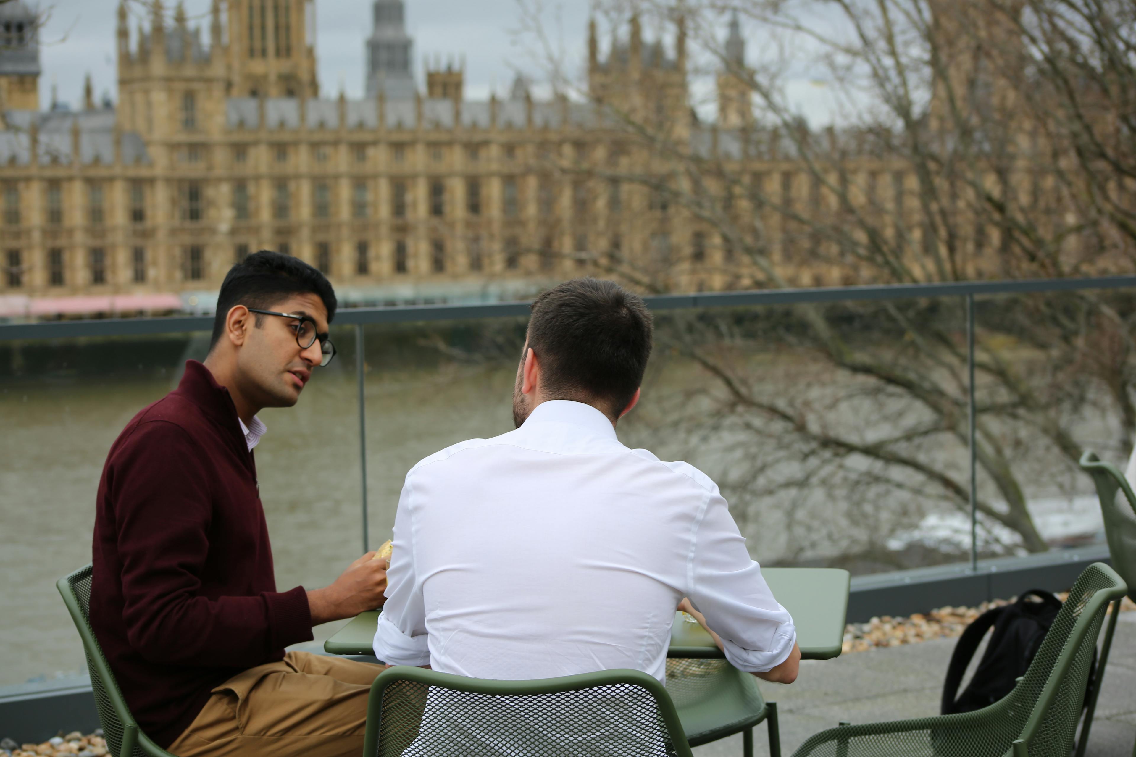 People chatting at a table sat on a balcony overlooking the Thames and Houses of Parliament