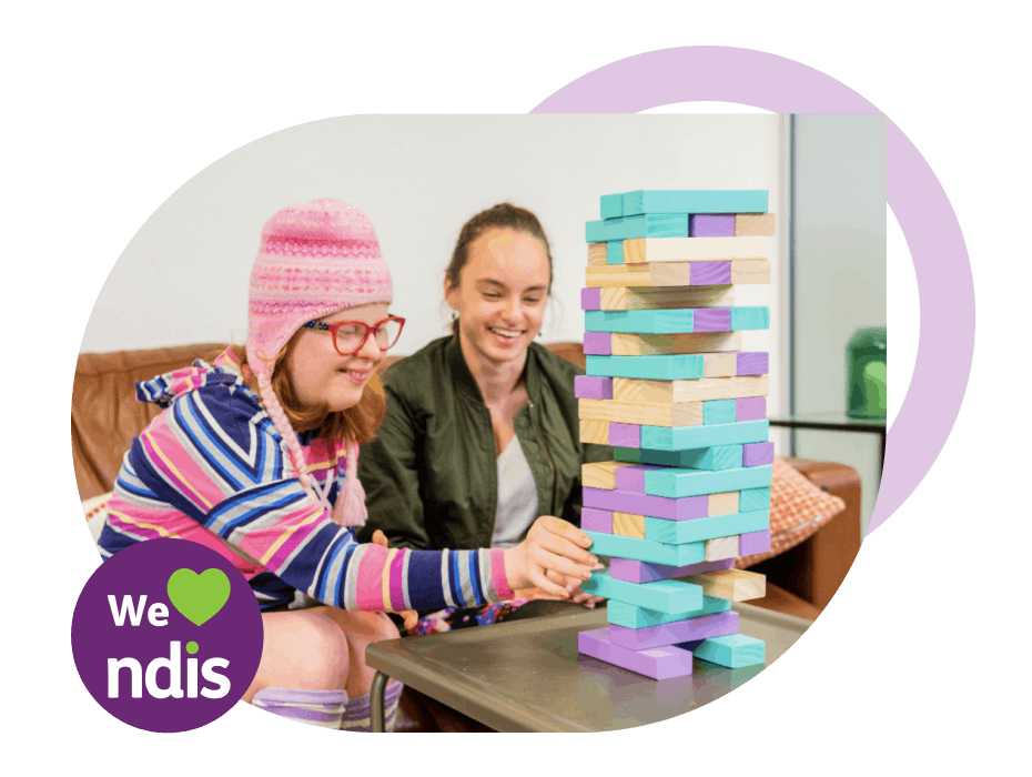 A Like Family support worker and Member playing Jenga with large colourful blocks. A purple "We heart NDIS" logo is to their left.