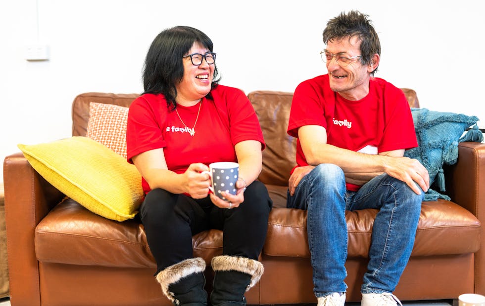 A man and a woman in red Like Family t-shirts sitting on a brown leather couch, looking at each other