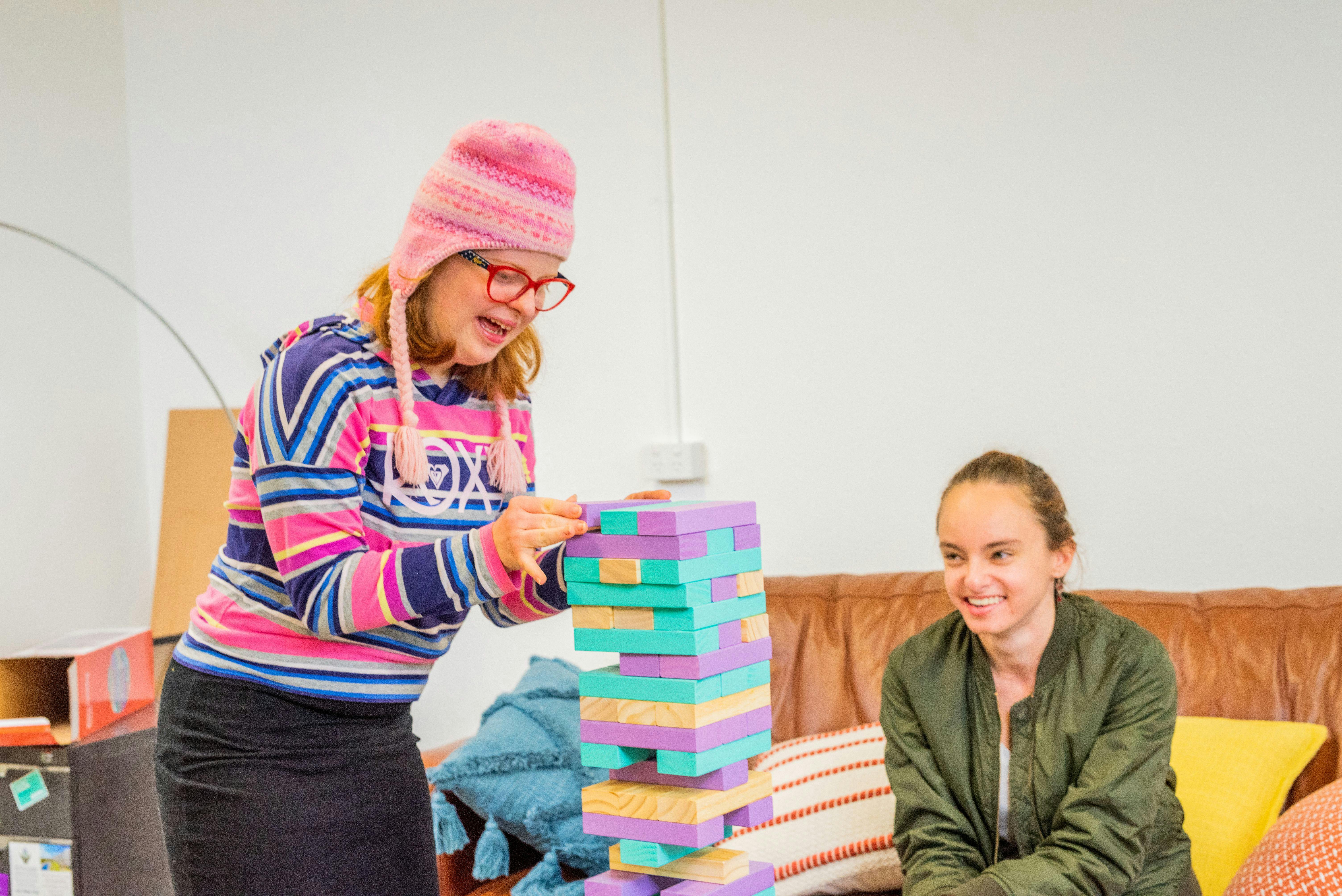 A Member places a Jenga block at the top of a colourful tower of blocks, while her Social Carer watches