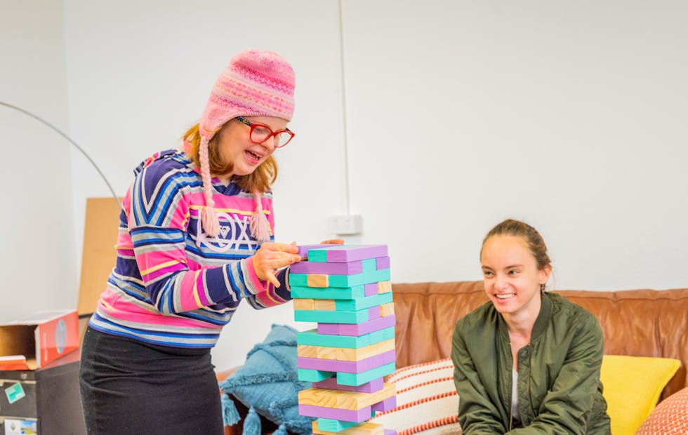 A Member places a Jenga block at the top of a colourful tower of blocks, while her Social Carer watches