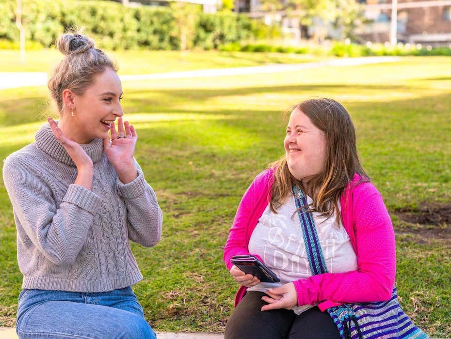 A young woman holding her hands up to her face and smiling to another woman who is laughing.