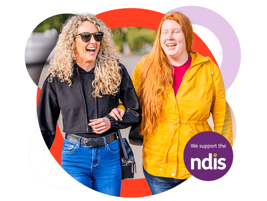 A Like Family member and her Social Carer walk along the street, arms linked. The woman on the left wears sunglasses and a black hoodie. The woman on the right wears a yellow jacket. A "We support the NDIS" sticker is on the right.