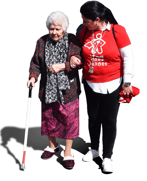 A female Social Carer holding the arm of an elderly woman, helping her to walk while turning to face her while saying something. The elderly woman has a walking stick.
