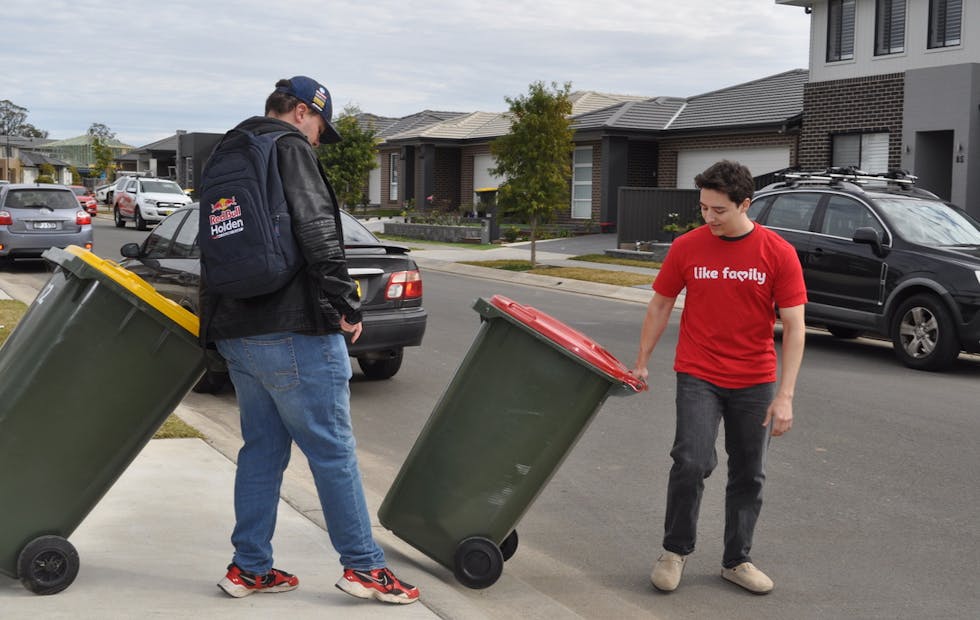 Two men taking out the bins. One is wearing a red Like Family shirt