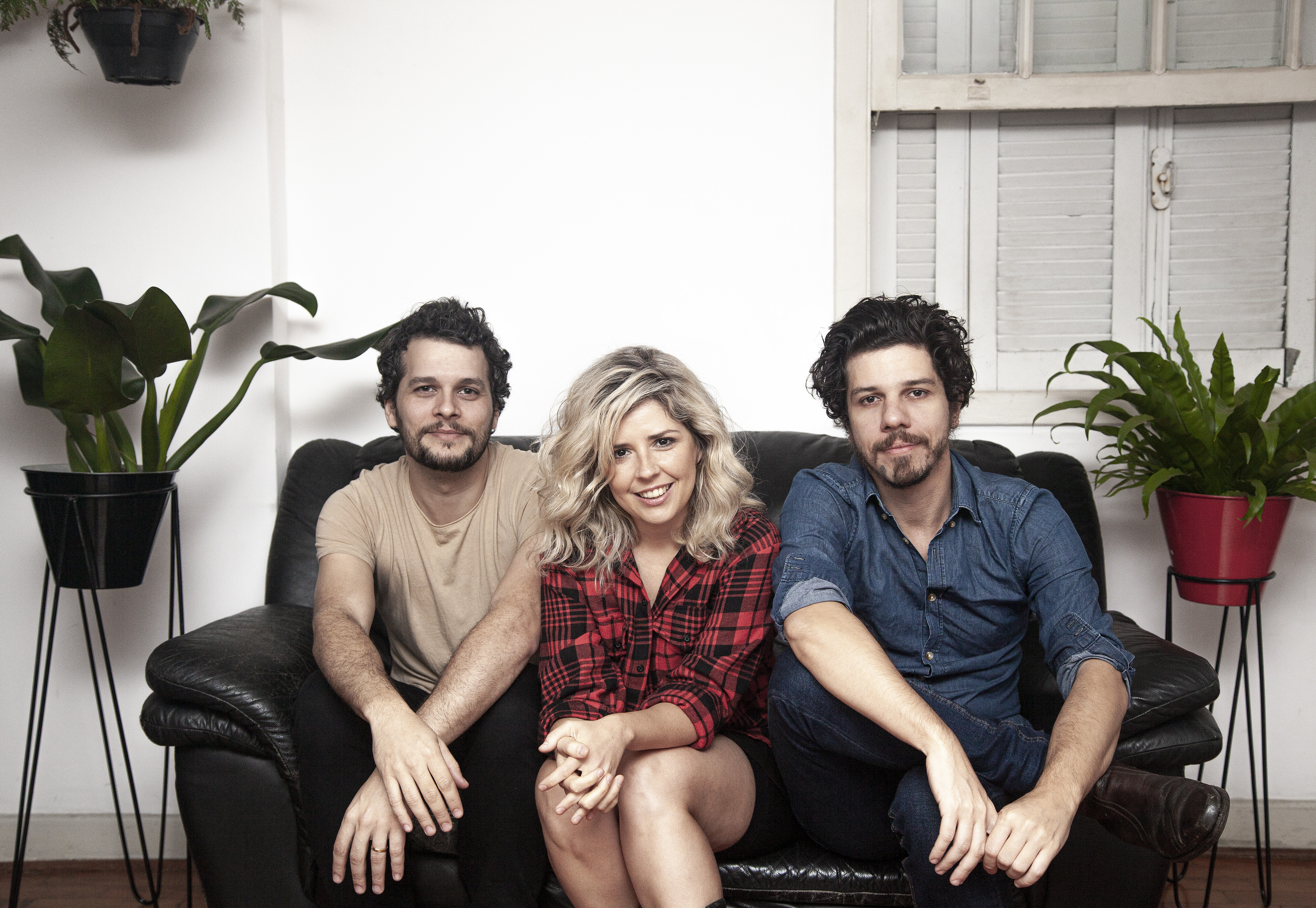 Reginaldo, Fernanda e Helio sentados em um sofá em frente a uma janela. Dos dois lados, há jarros de plantas.