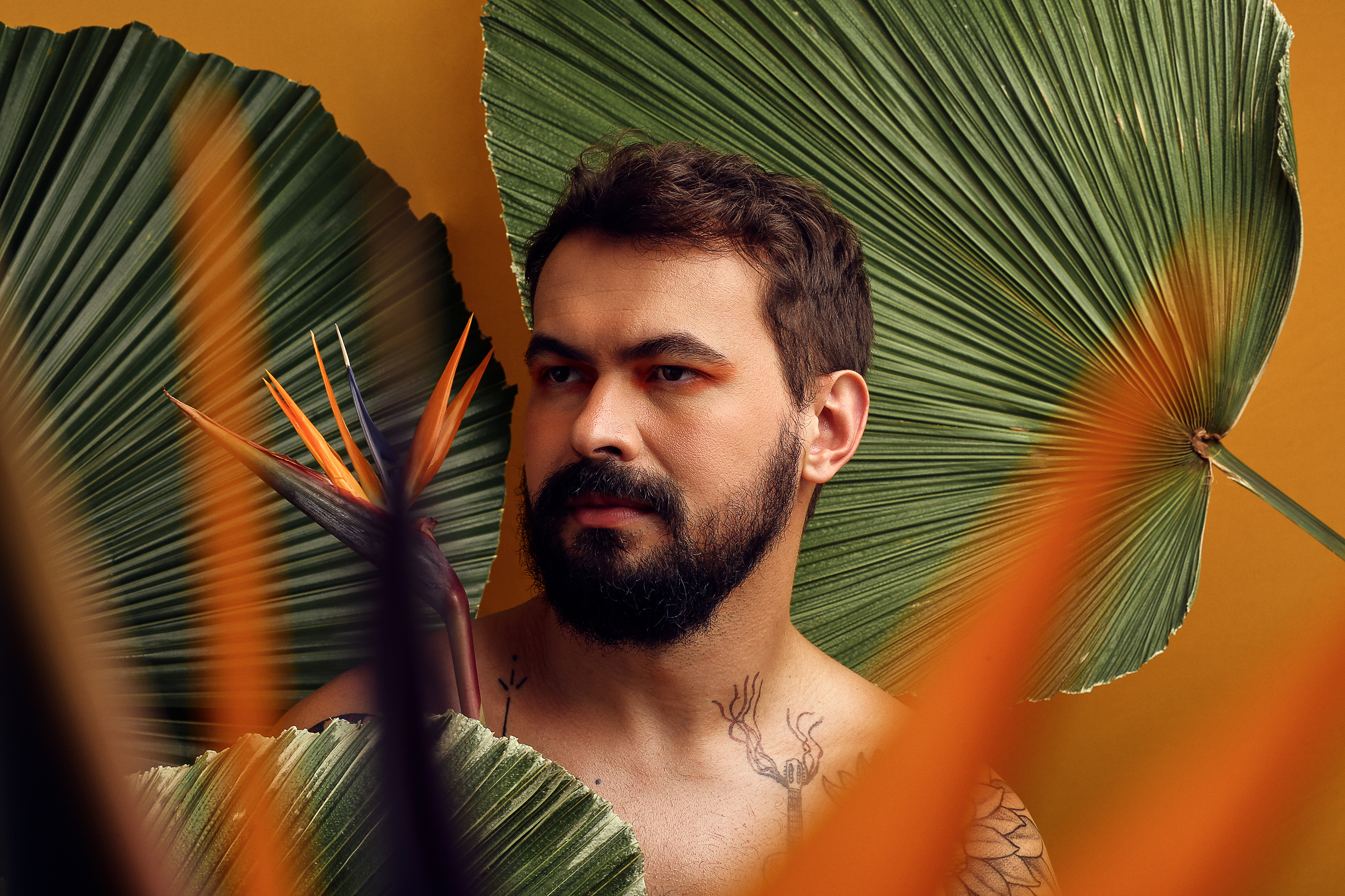 Bemti aparece do ombro para cima, sem camisa e com folhagens ao redor. A parede, ao fundo, é amarela. Ele olha para o lado.