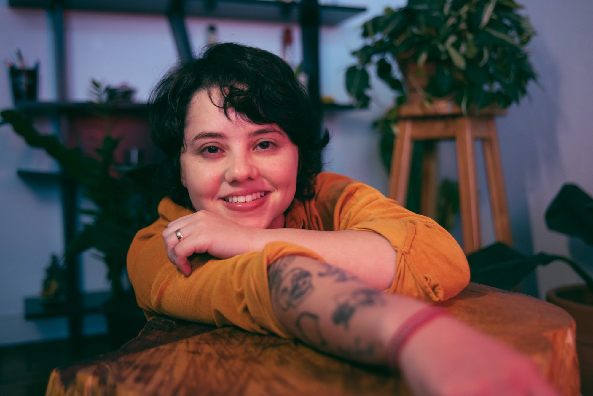 Ana Müller posa para a câmera. Apoiada em um de seus braços, está com a outra mão perto do rosto. Ana veste uma blusa amarela de mangas,seu antebraço está à mostra