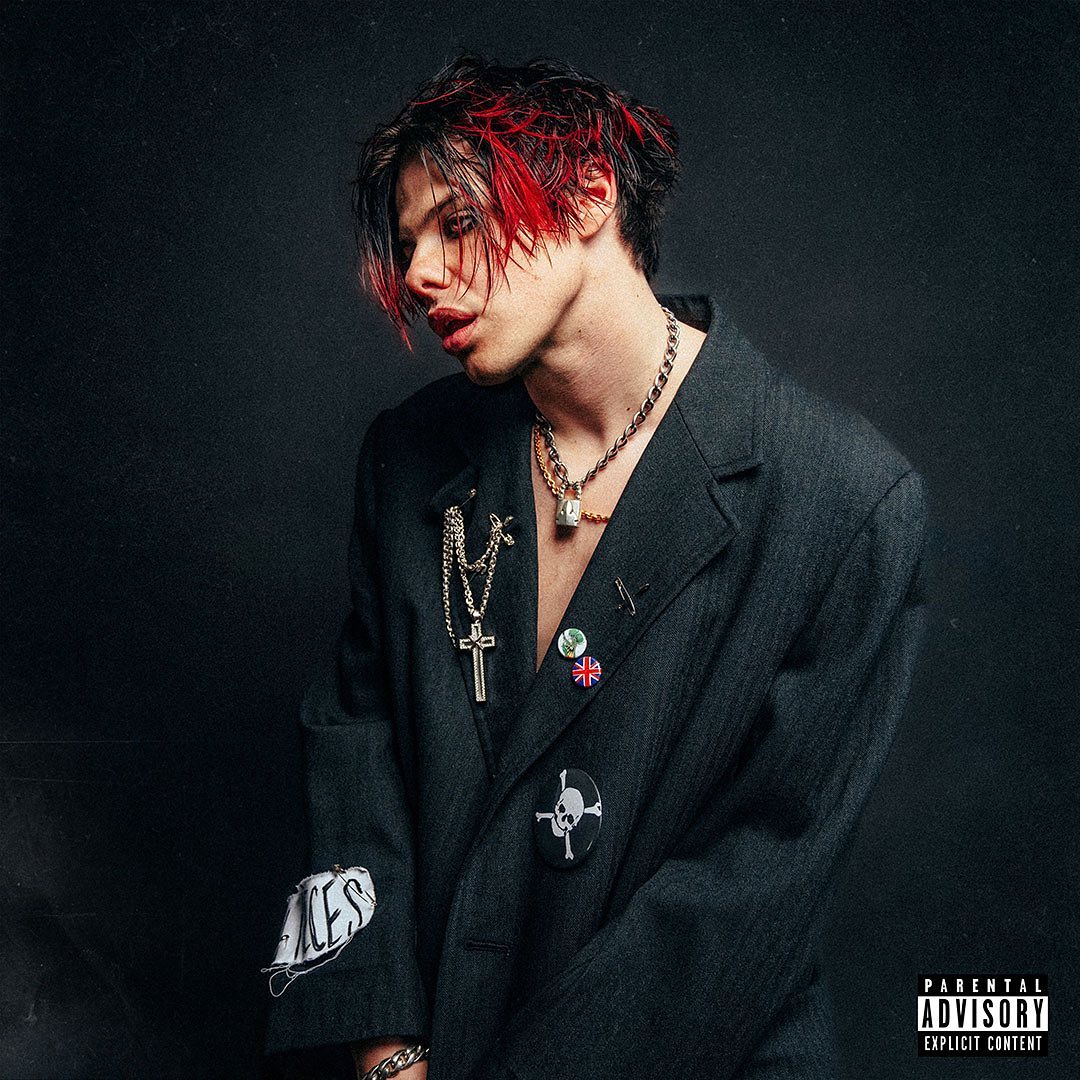 Yungblud é branco, tem o cabelo preto com as pontas vermelhas, que caem parcialmente sobre o rosto. Veste apenas um blazer, com alguns bottons e uma corrente pesada no pescoço.