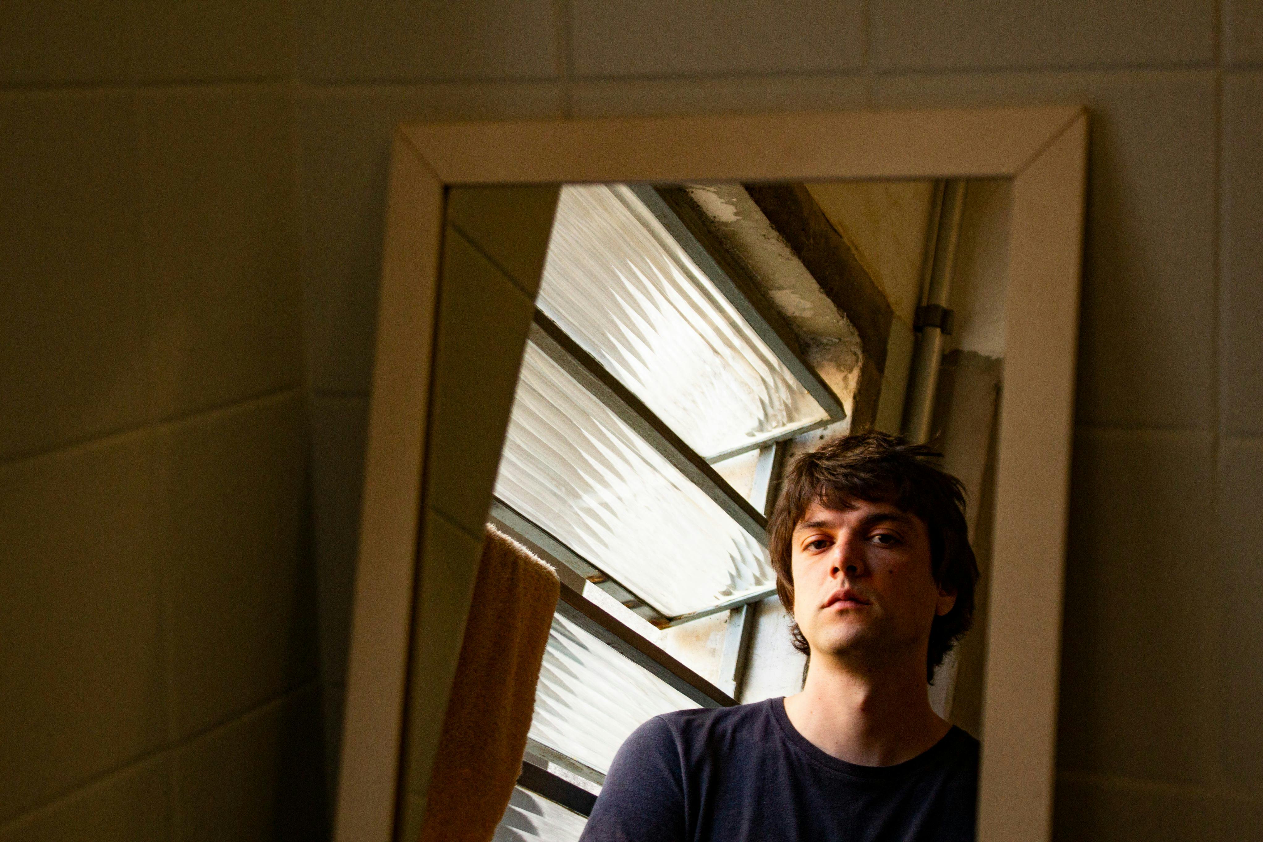 Juliano fm Costa aparece encarando o espelho. Atrás do acessório, há a parede, igualmente bege. A janela, atrás de Juliano, aparece no espelho.