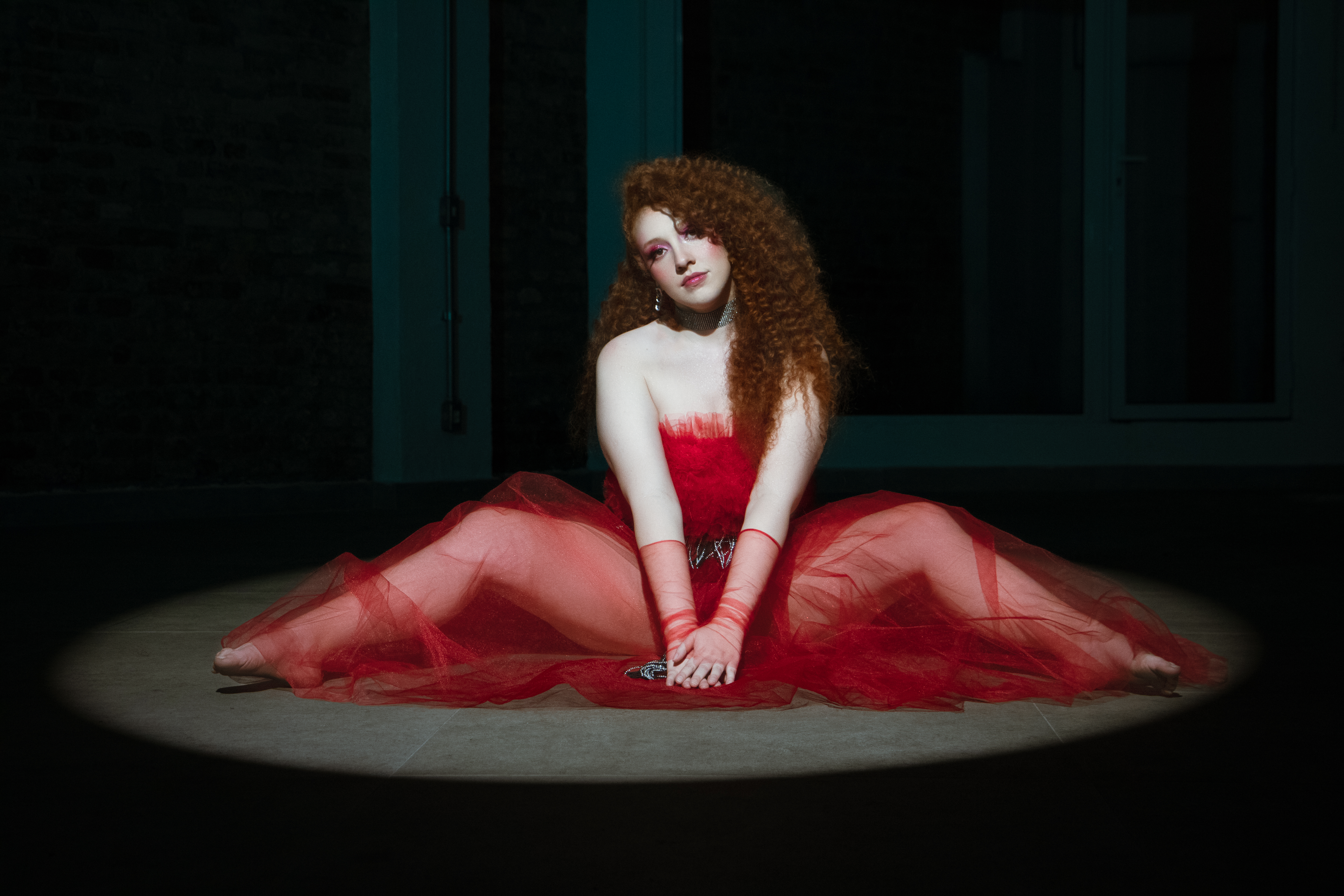 Bea Duarte está sentada no chão, de vestido vermelho. Ela tem o cabelo ruivo cacheado.