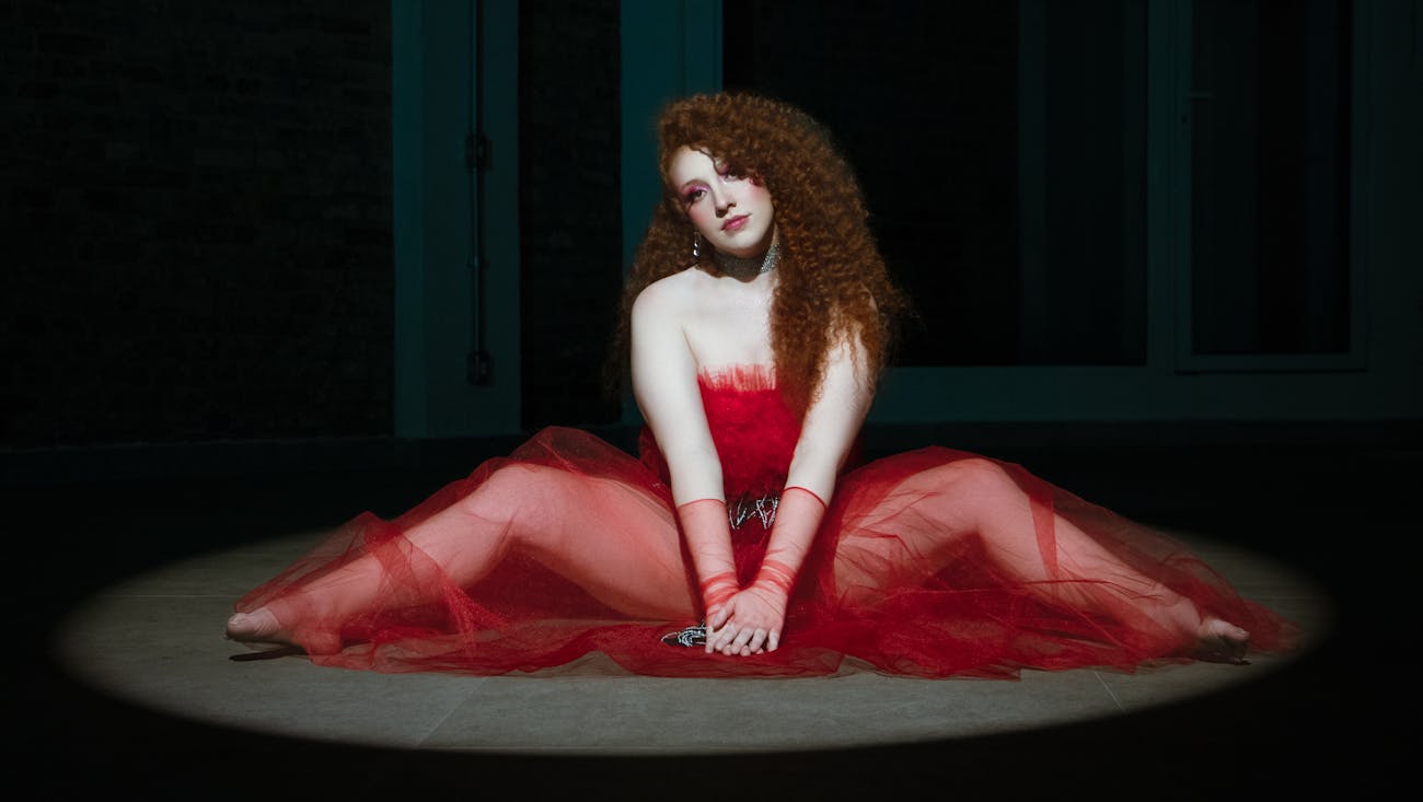 Bea Duarte está sentada no chão, de vestido vermelho. Ela tem o cabelo ruivo cacheado.