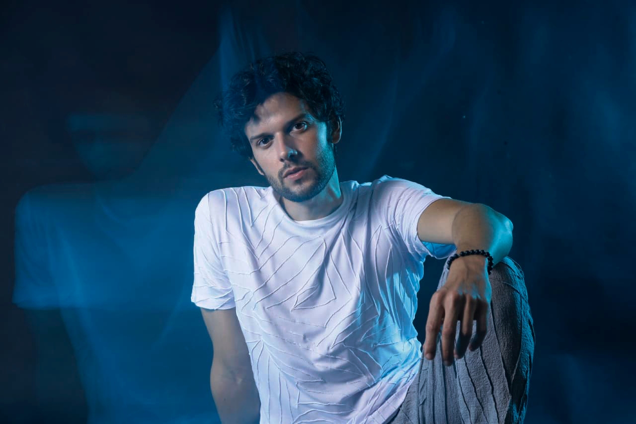 Davi Cartaxo está sentado, com uma perna levantada, onde apoia o braço esquerdo, olhando para a câmera. O cantor está inteiro de camiseta branca na frente de um fundo azul. Há um reflexo dele aí fundo.