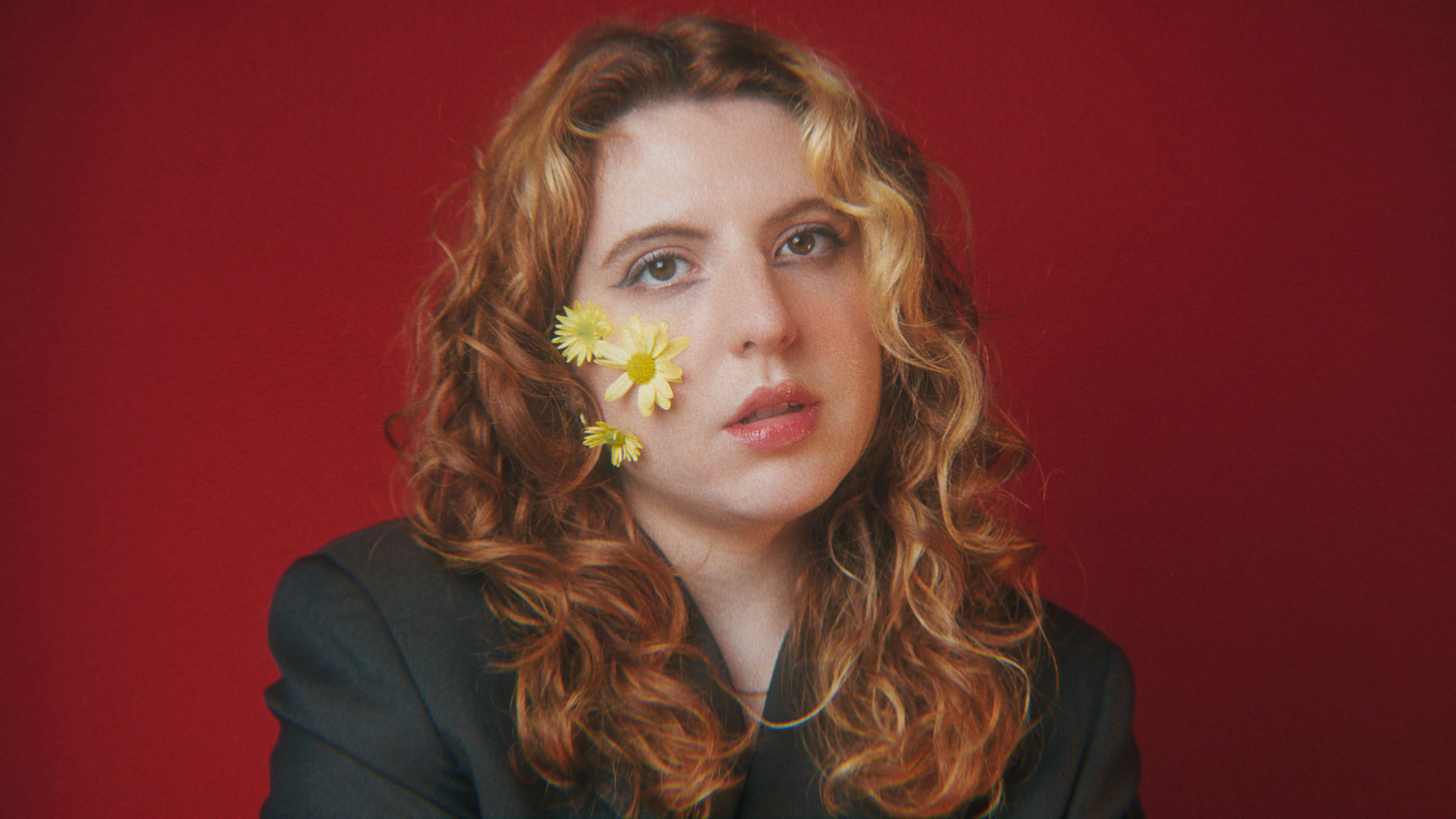 Tuany é uma mulher branca e loira. Ela está a frente de um fundo vermelho e tem flores amarelas colocadas no rosto. 