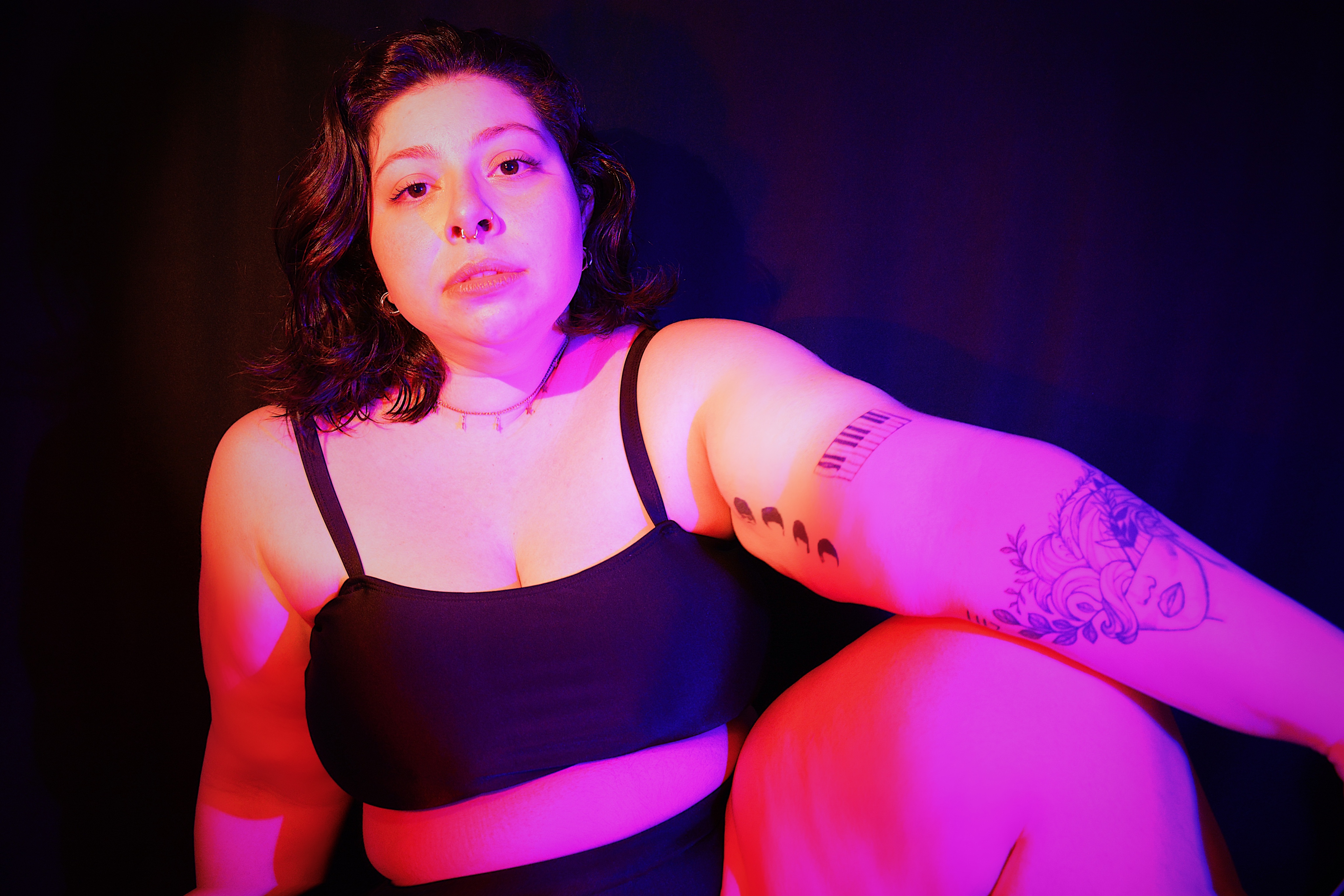 Paula Azzam está de cropped preto, deitada quase que de lado, olhando fixamente para a câmera. Refletidas nela, há luzes cor de rosa.