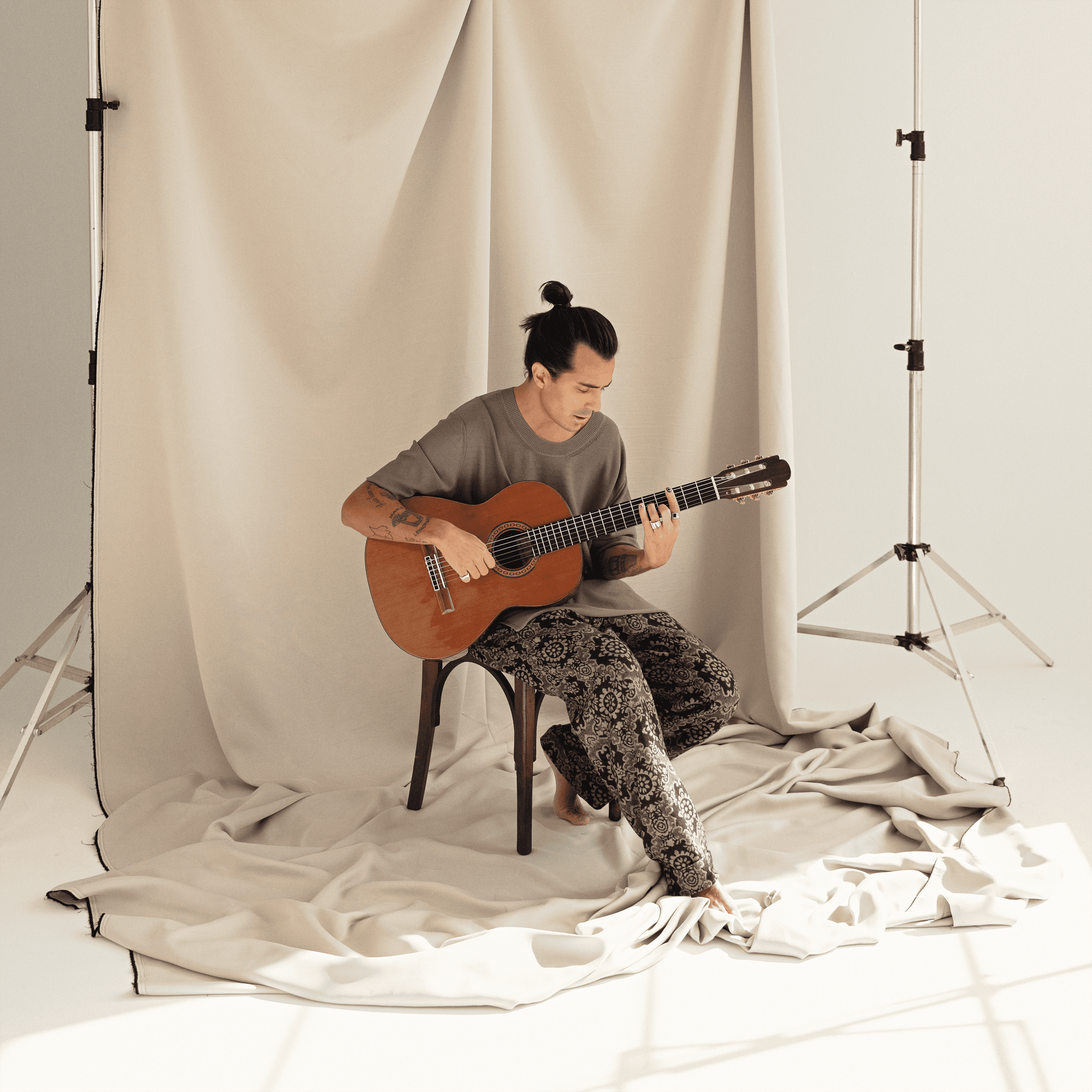 Pe Lu aparece a frente de um pano bege, em um estúdio. Ele está sentado em uma cadeira, com roupas largas e o cabelo preso. Tocando violão, ele olha para o instrumento. A foto foi tirada de longe.