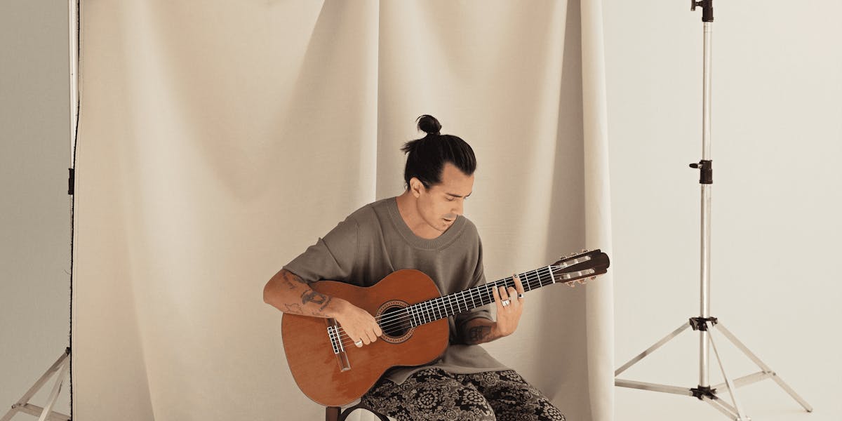 Pe Lu aparece a frente de um pano bege, em um estúdio. Ele está sentado em uma cadeira, com roupas largas e o cabelo preso. Tocando violão, ele olha para o instrumento. A foto foi tirada de longe.