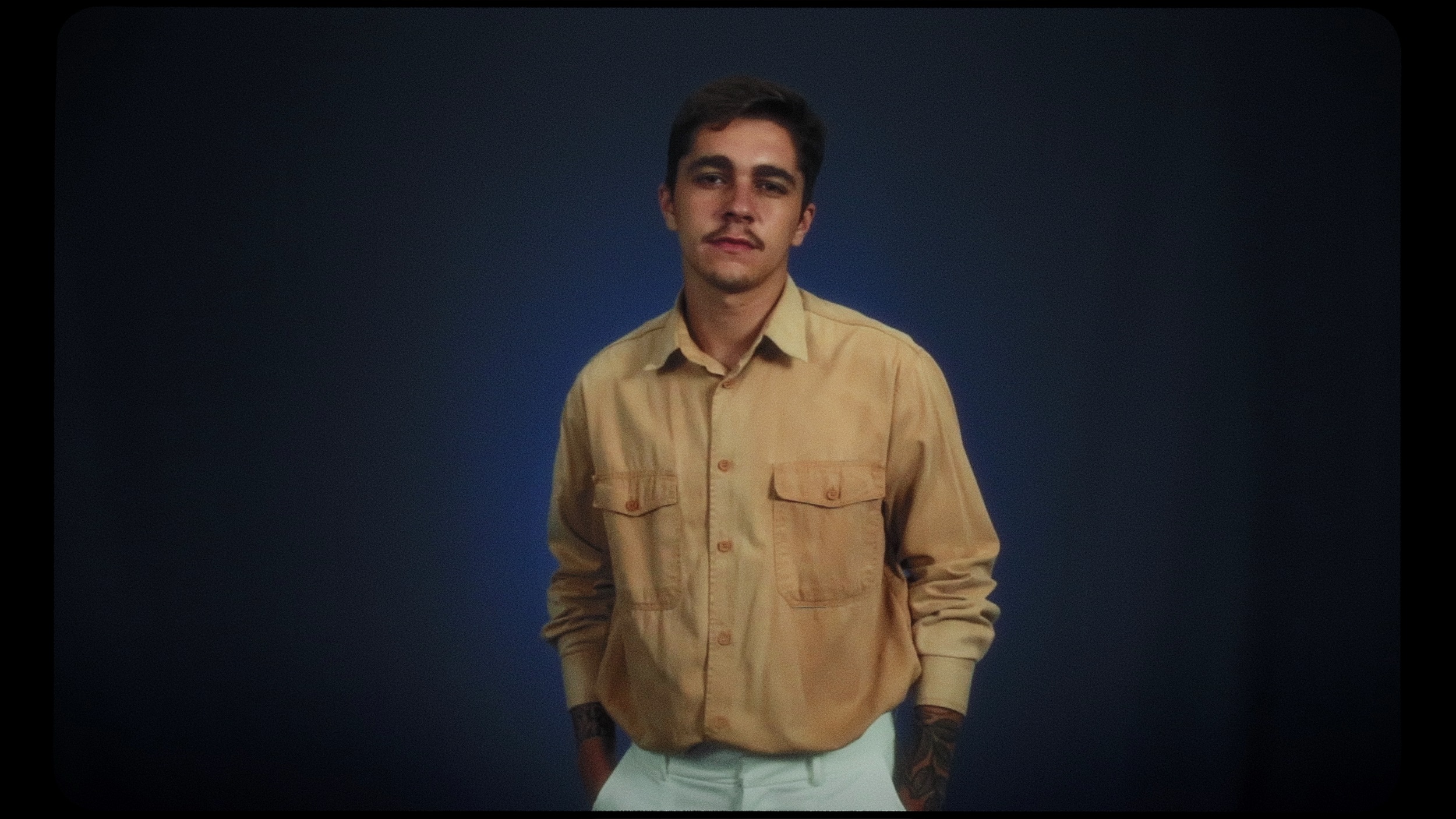 Sávio está a frente de um lençol branco, com uma blusa bege e uma calça branca. 