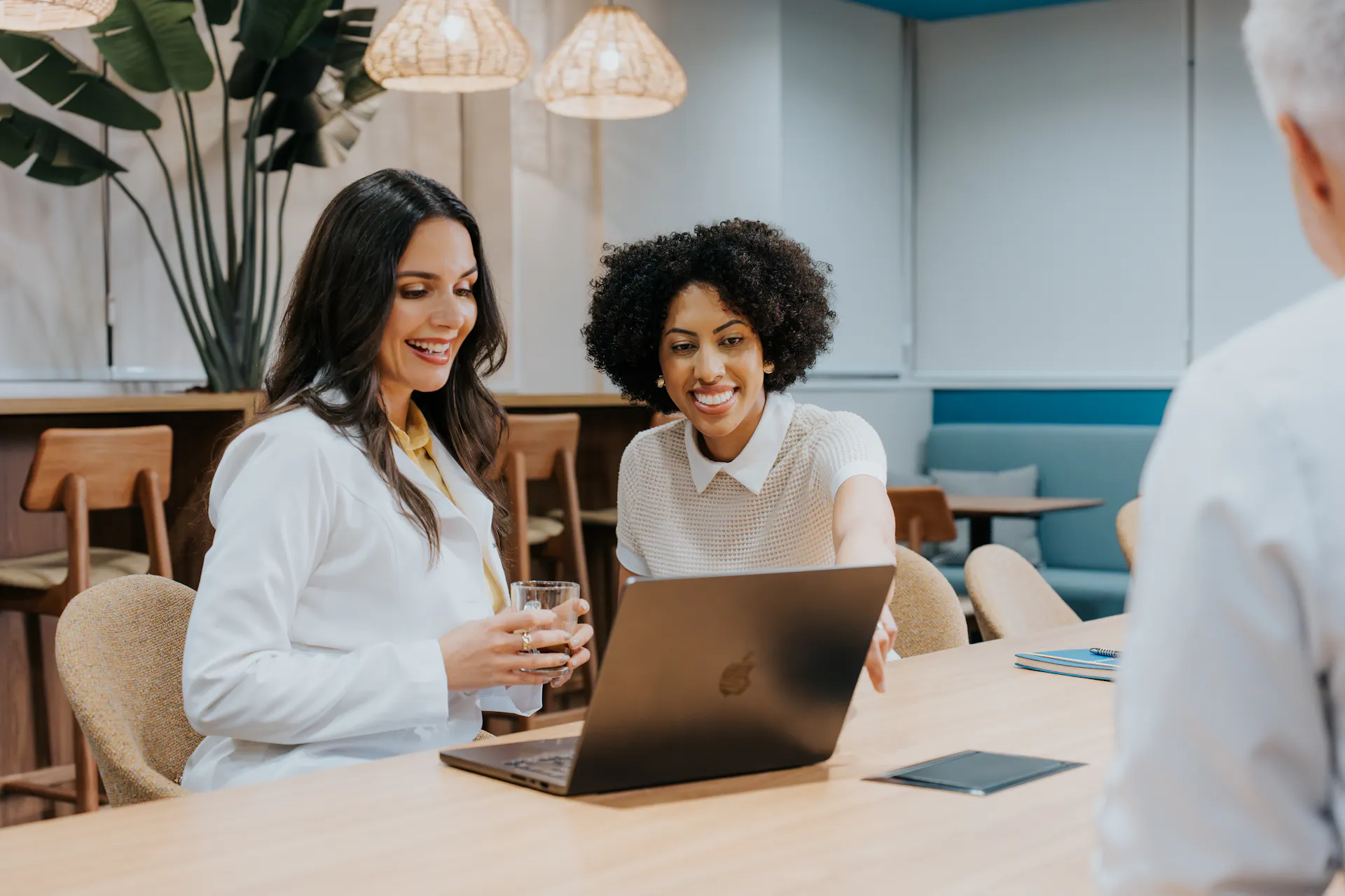 Profissional de saúde de jaleco branco e uma mulher sentadas à mesa em um ambiente moderno de coworking, observando um notebook enquanto conversam com outra pessoa à frente. A profissional de saúde segura um copo de água e sorri, enquanto a outra mulher aponta para a tela do computador. O espaço é iluminado por luminárias pendentes e conta com decoração em madeira e plantas, transmitindo um ambiente acolhedor e colaborativo.