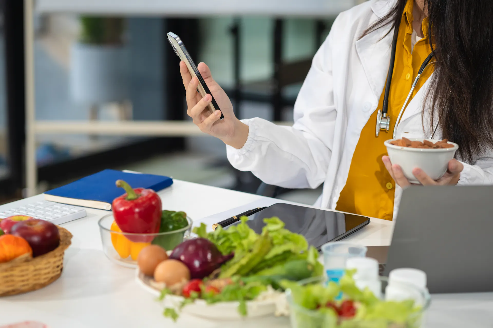 Nutricionista utilizando o celular no consultório ao lado de alimentos saudáveis e notebook.
