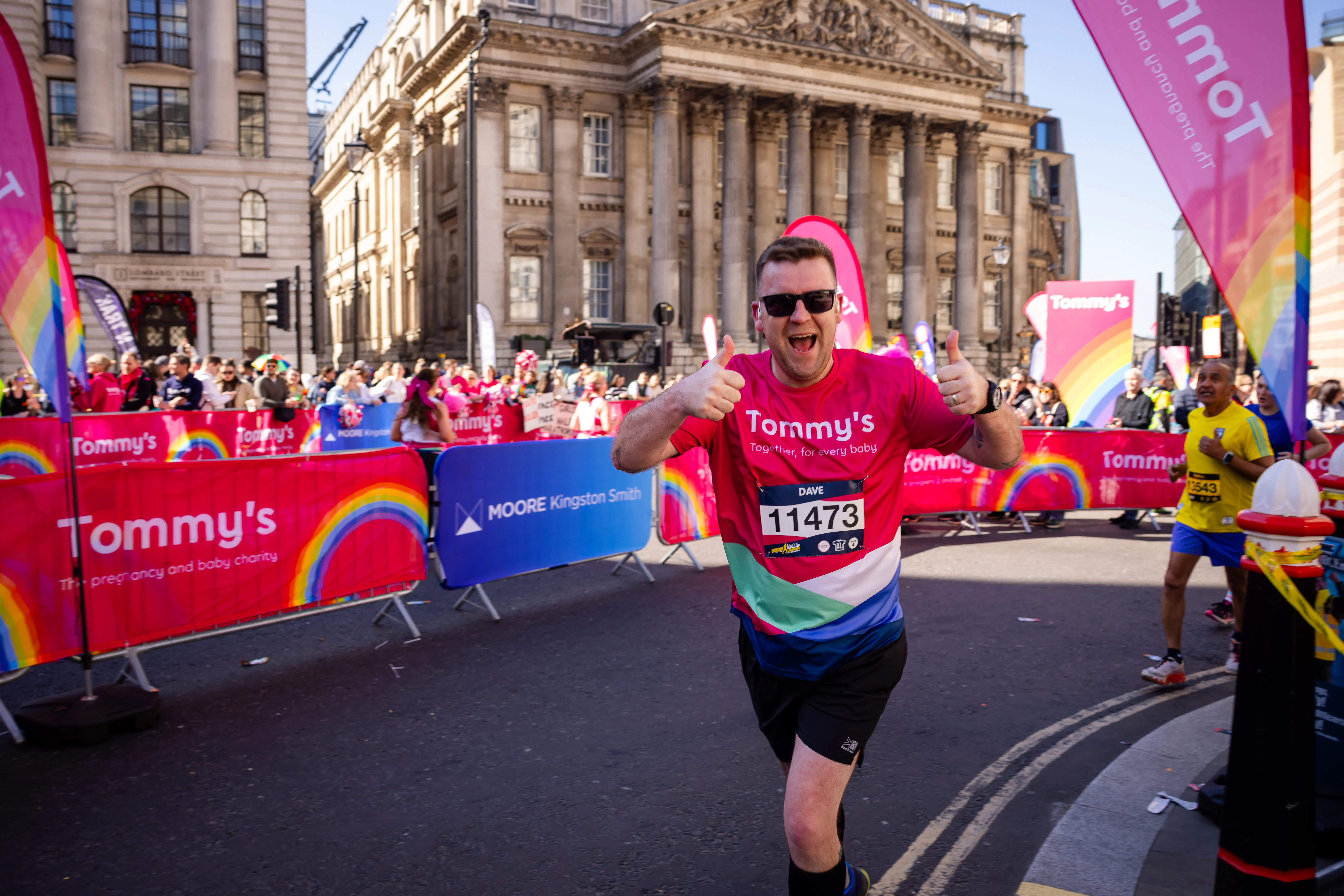 Runner in a Tommy’s charity shirt giving two thumbs up while taking part in a city centre road race, with spectators and rainbow-branded event banners in the background.