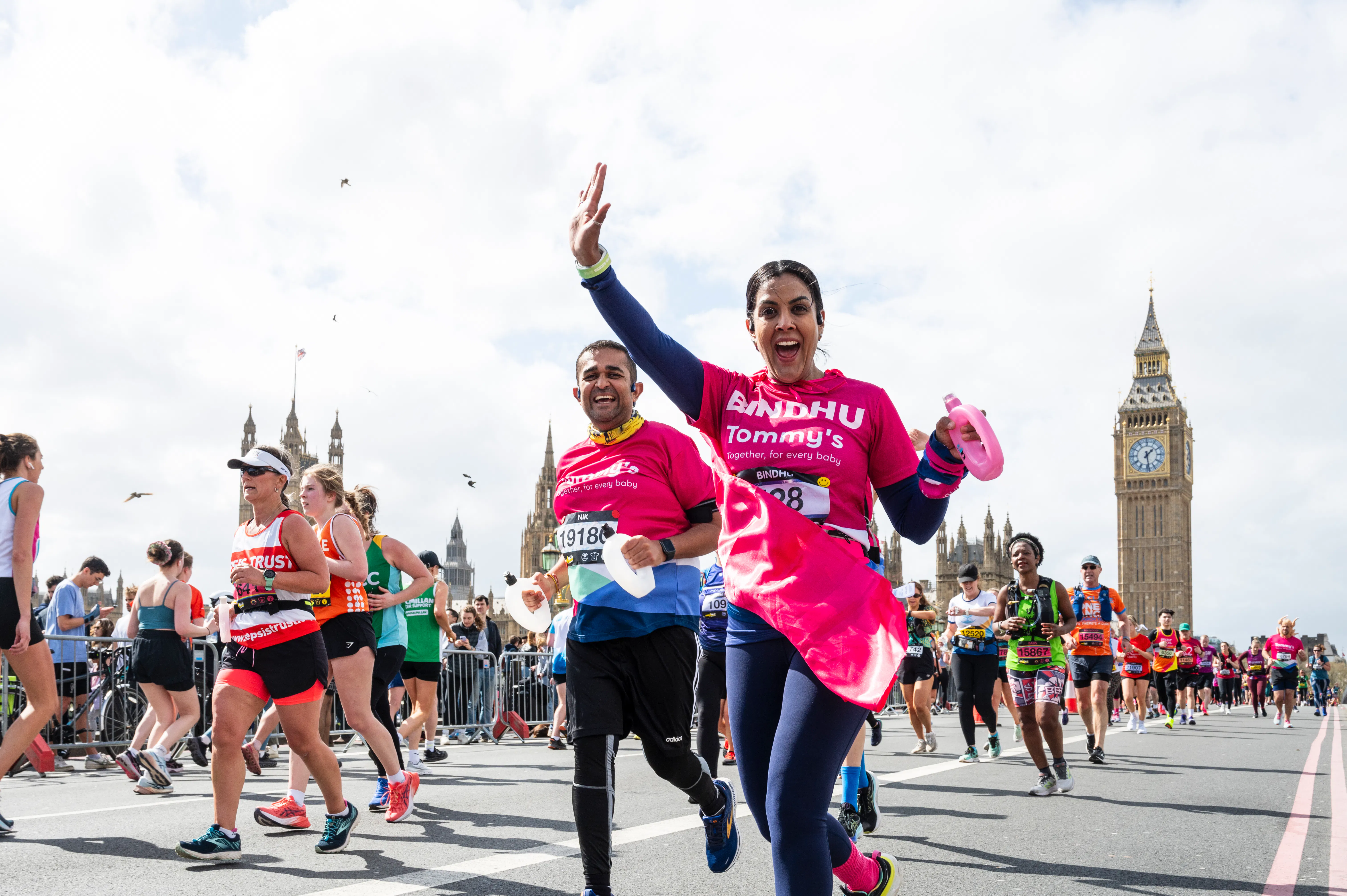 Runner in a bright pink charity shirt waves while taking part in a road race near Big Ben, surrounded by fellow participants.