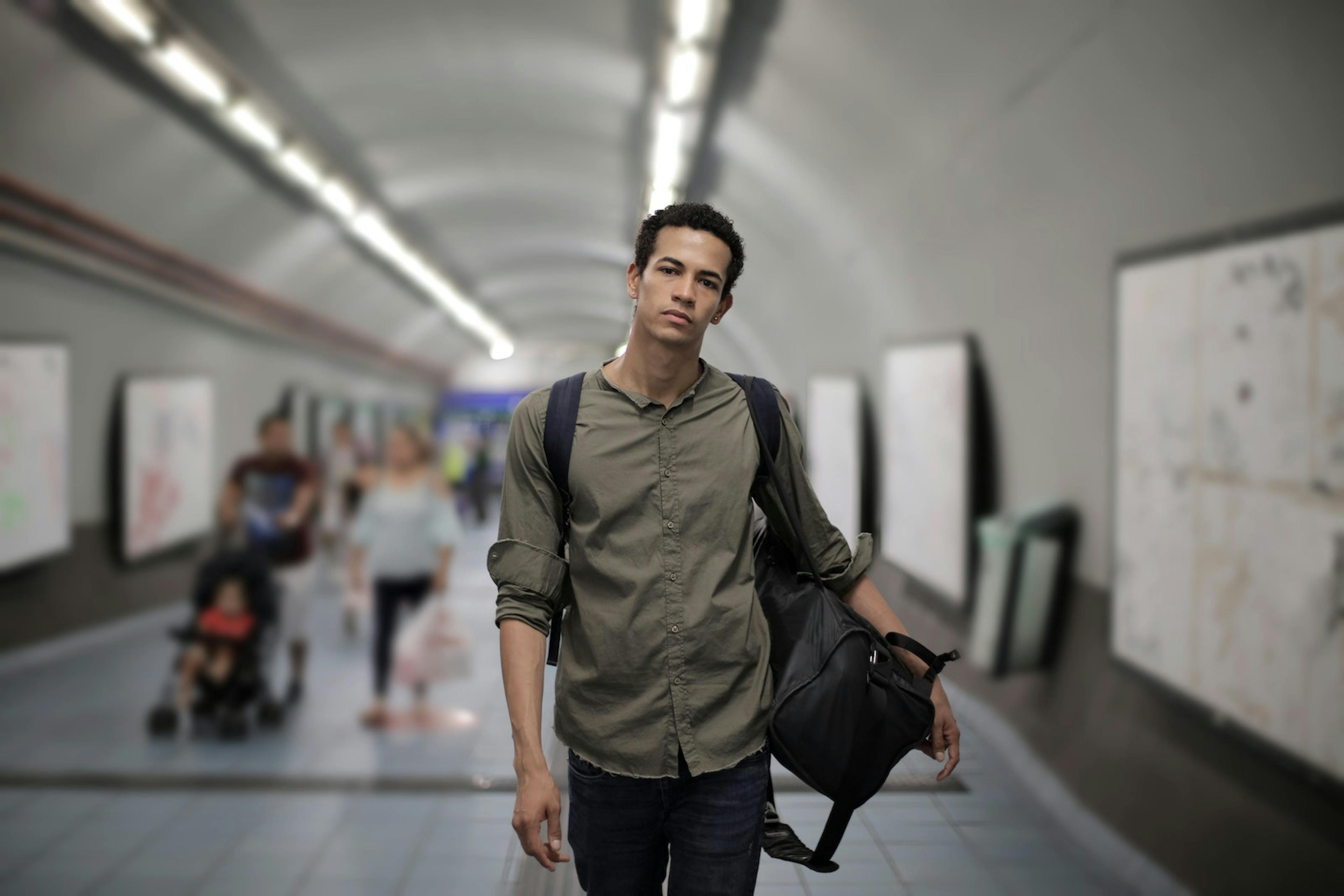 a young, sad looking man walks through a subway tunned while carrying a duffel bag