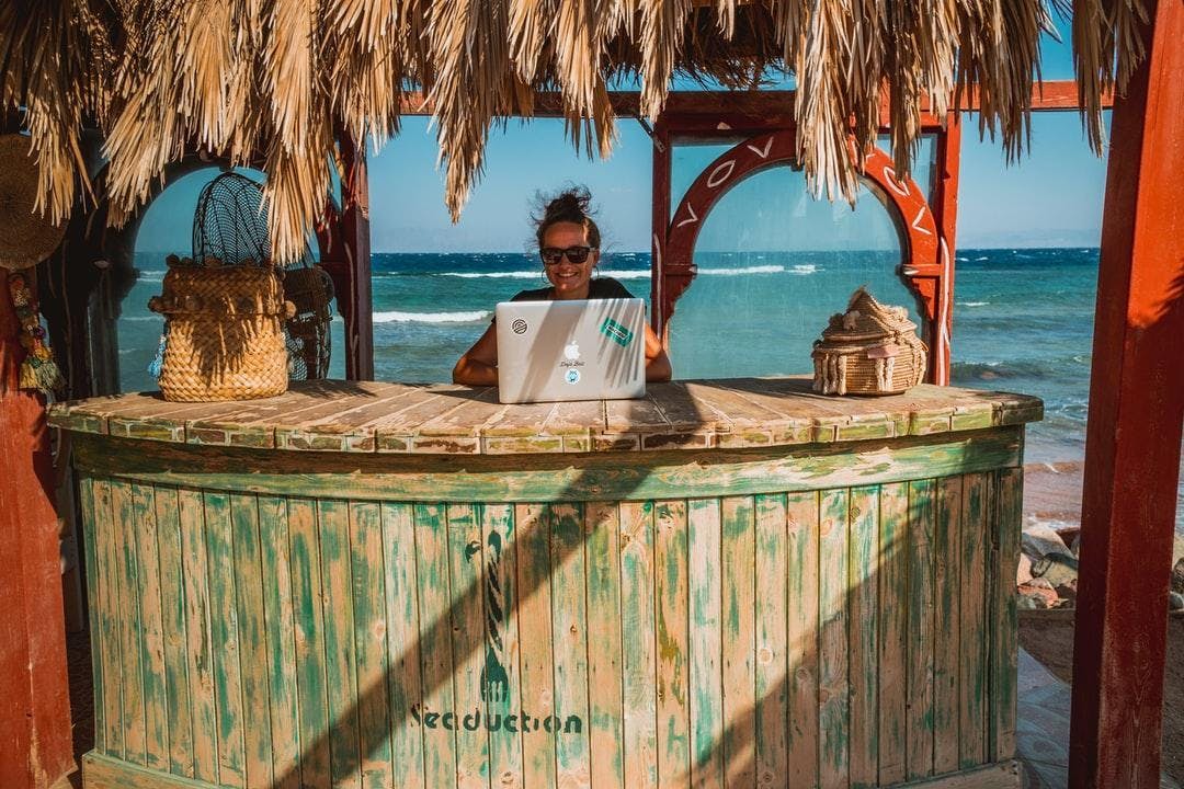 Worker sits in a cabana in front of the ocean while working on laptop.