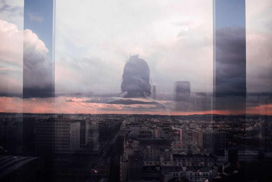 a woman looking out is reflected in the window of an upper story window, representing mental health needs of employees