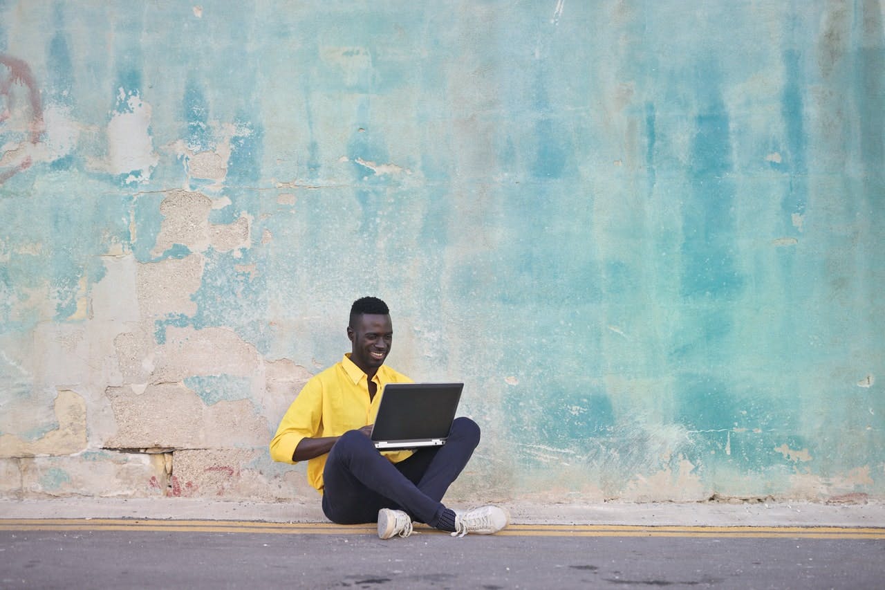 a young black man smiles while sitting on a curb in front of a light blue wall and working on his laptop