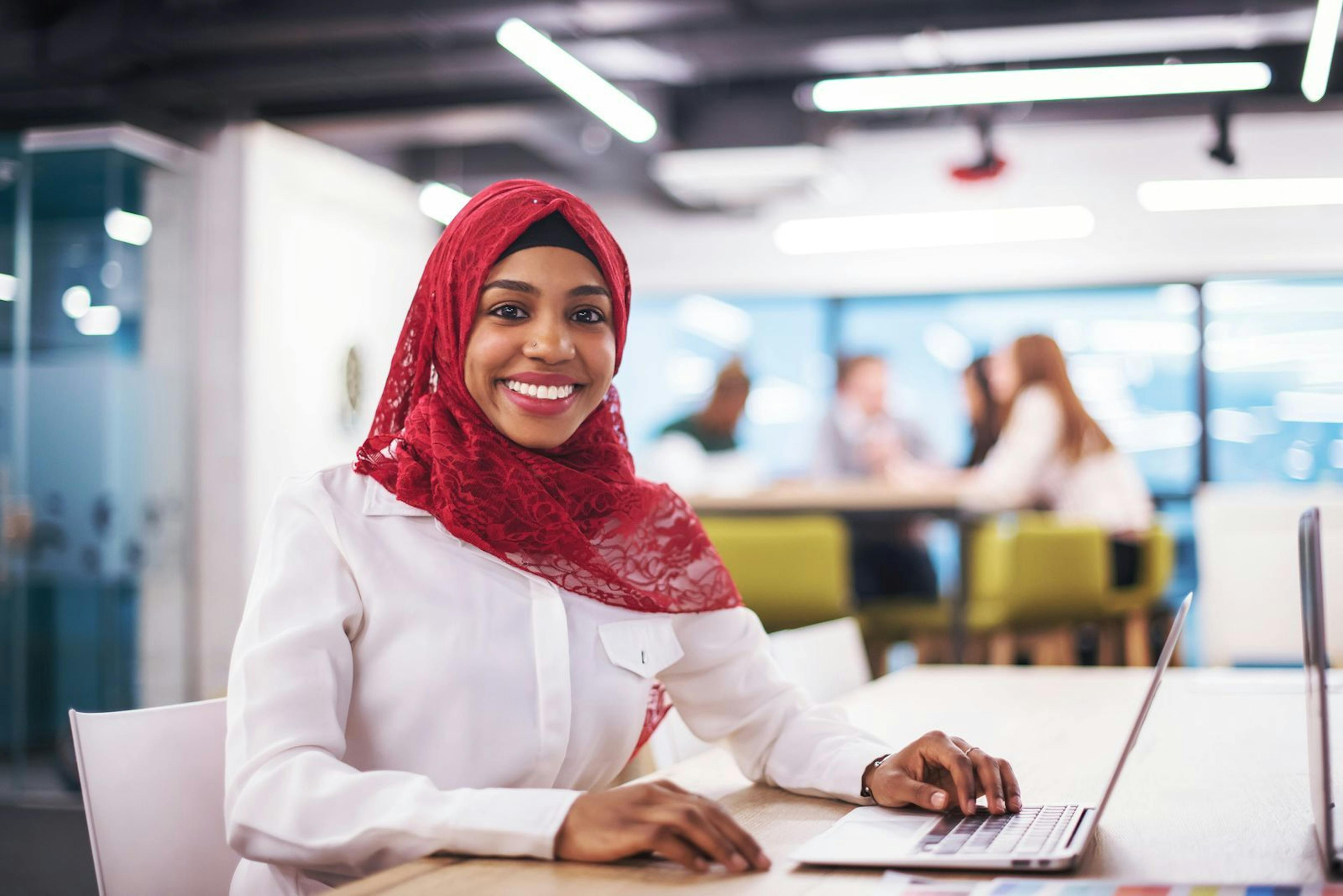 a smiling woman in a hijab working on how to help refugees from a laptop