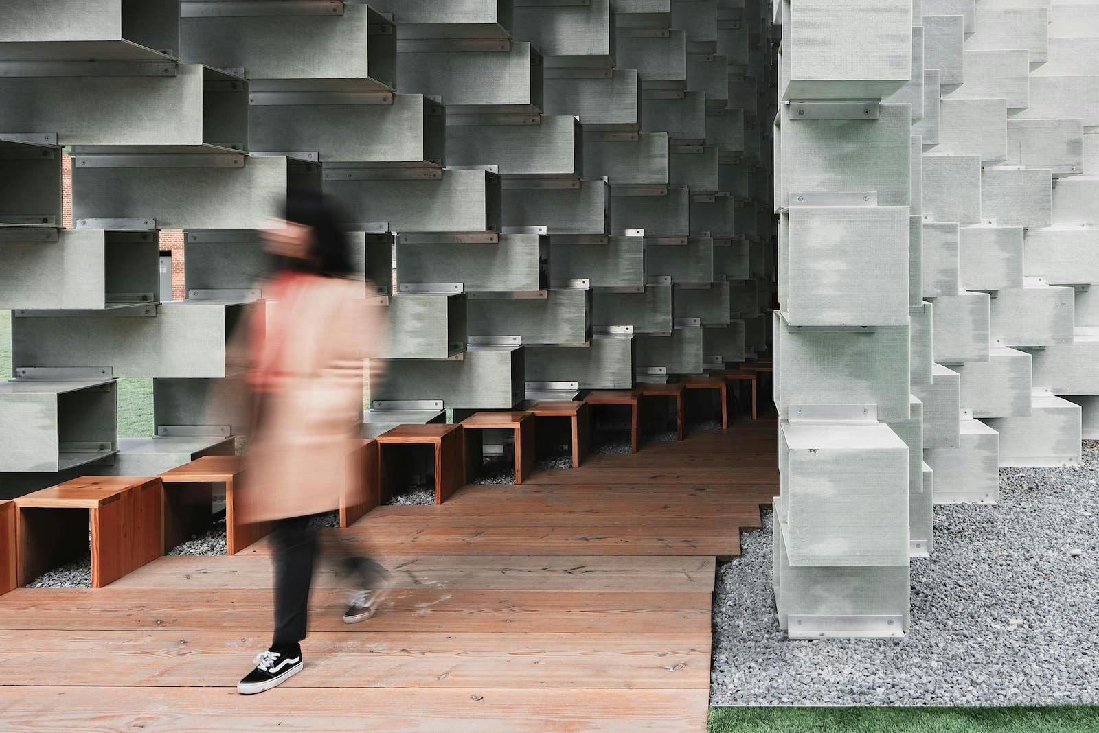 a woman walks on a wooden pathway with a metal sculpture wall behind her