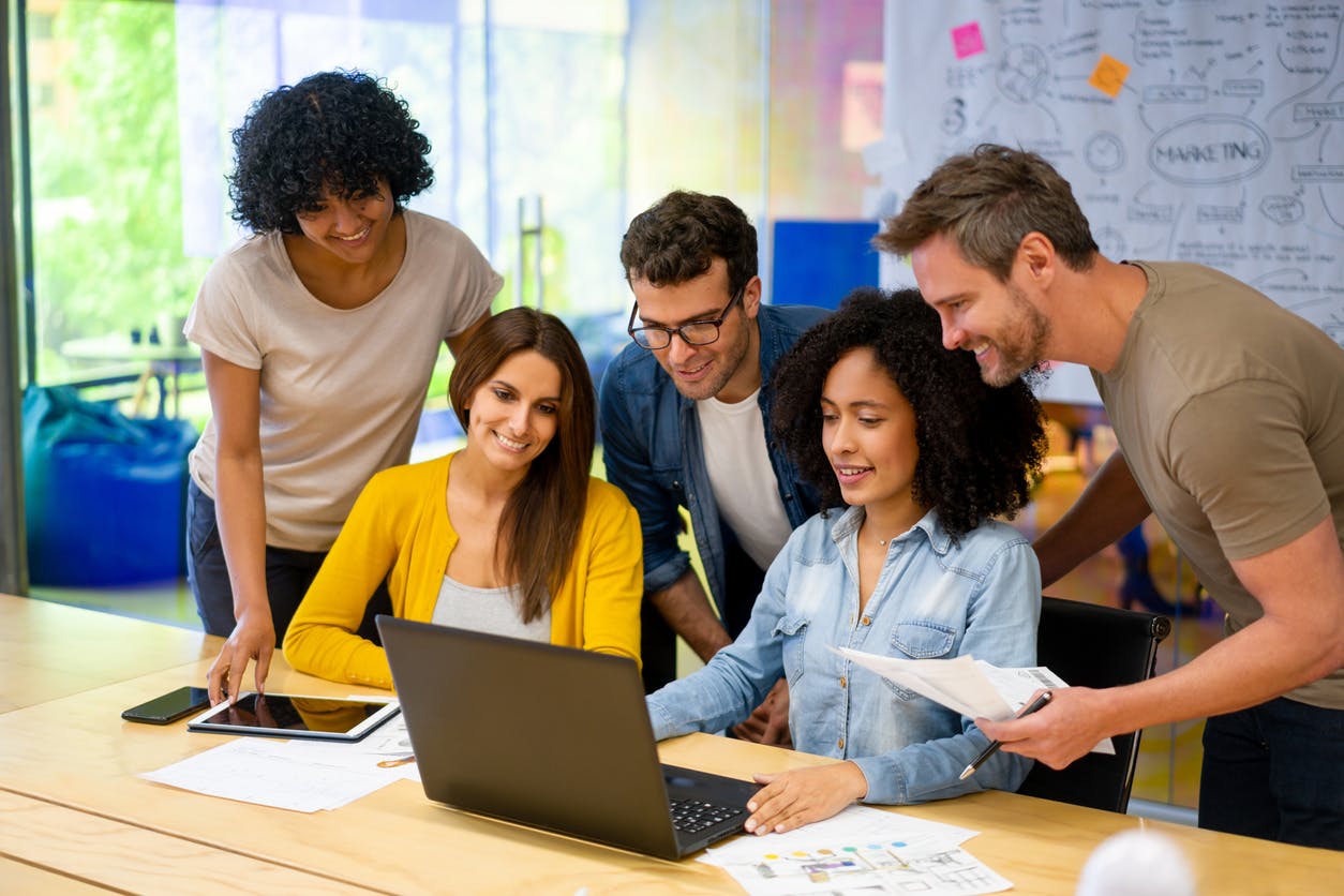 a group of five diverse coworkers gathered around a laptop, smiling while they work on a distributed workforce plan