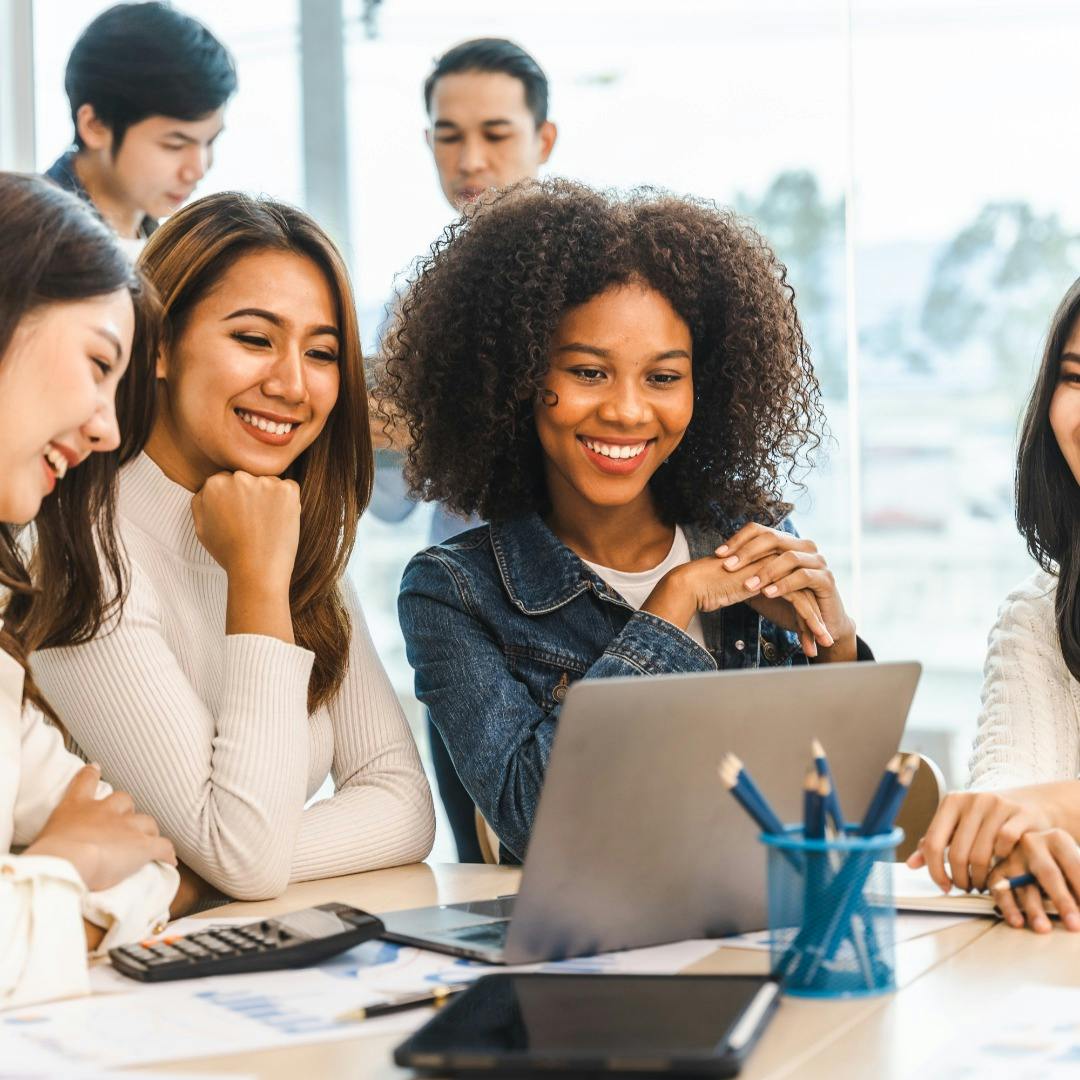 a group of divers women smile at a single laptop screen, presumably while on a video call