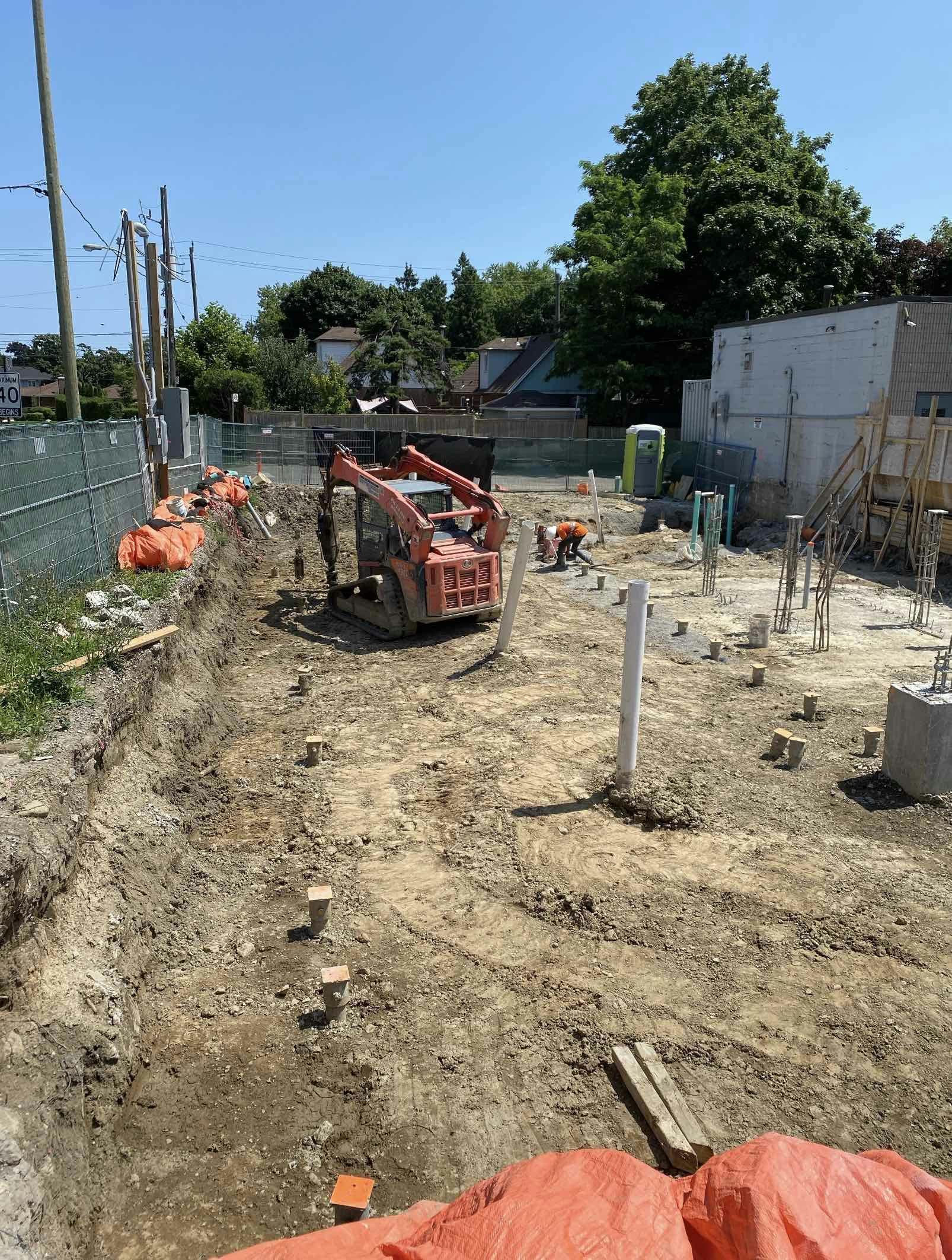 Construction site with an excavator, construction materials, and a worker in the background, surrounded by fencing and trees