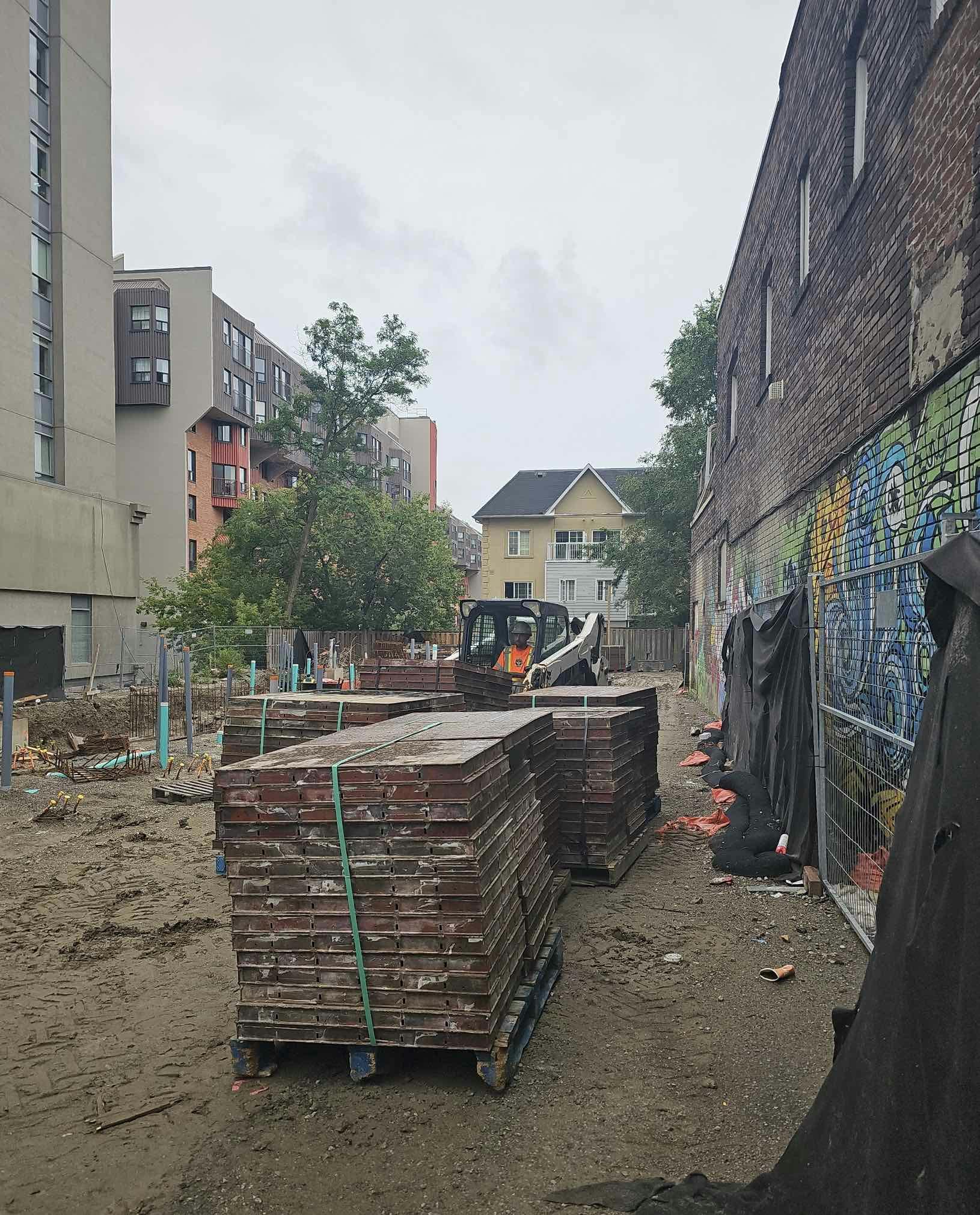 Construction site at 215 Wellesley St. with stacked building materials, a construction vehicle, and surrounding buildings