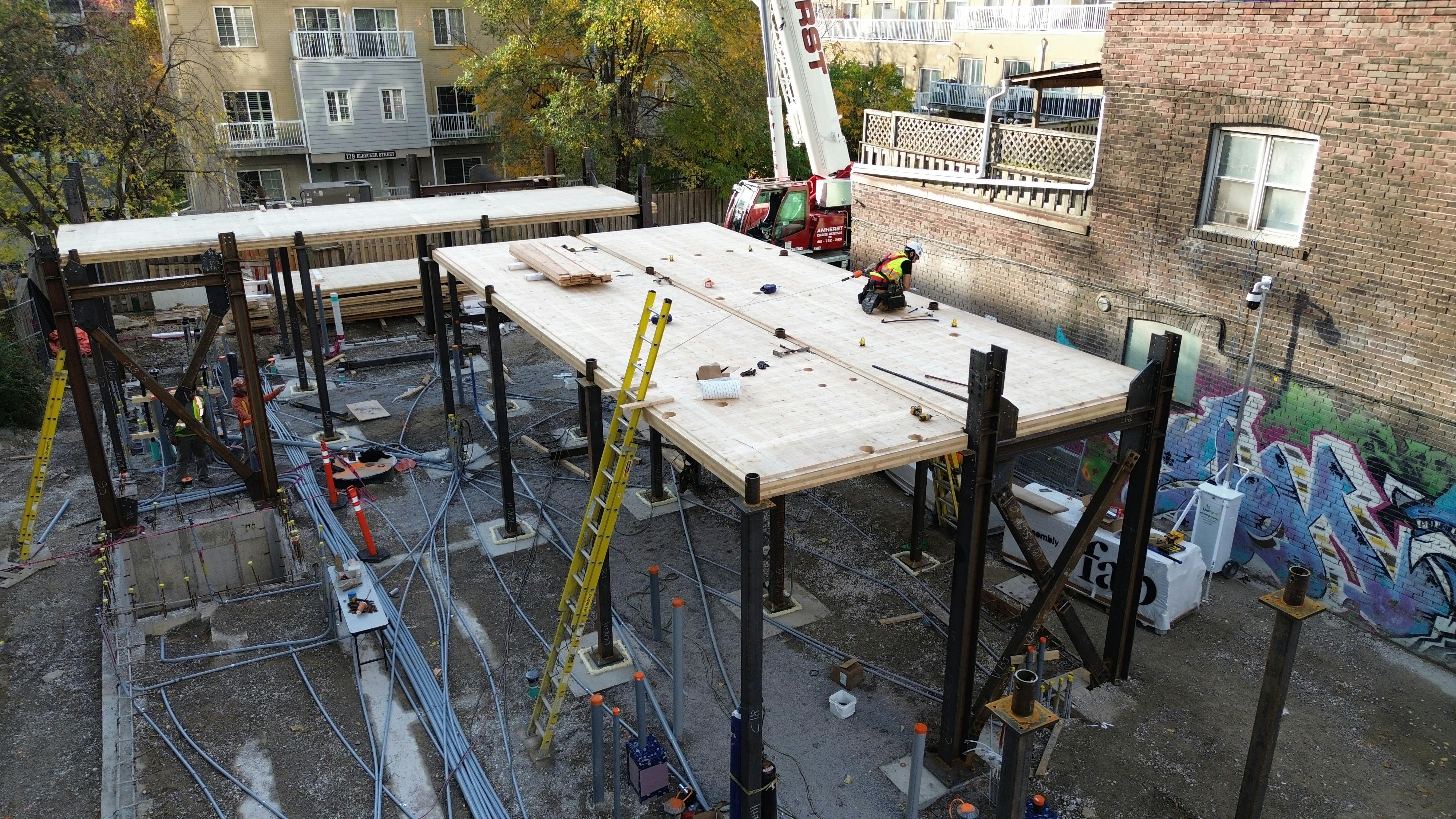 Construction site with mass timber platforms being installed, supported by steel beams anchored to concrete foundations. Electrical and plumbing conduits are visible below the structure, and a crane is positioned nearby.