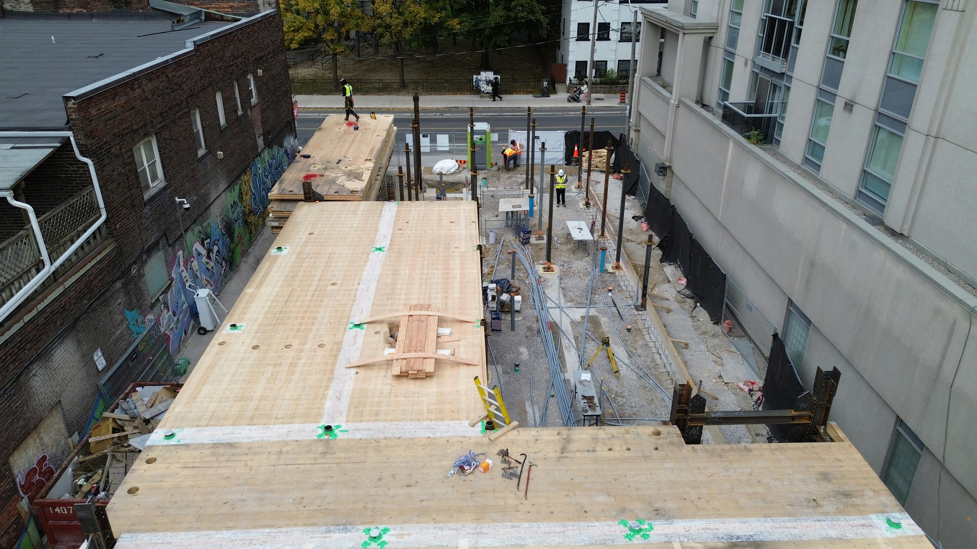 Aerial view of 215 Wellesley St showing two partially constructed wooden platforms supported by steel beams. Workers are spread across the site, some on the platform and others at ground level.