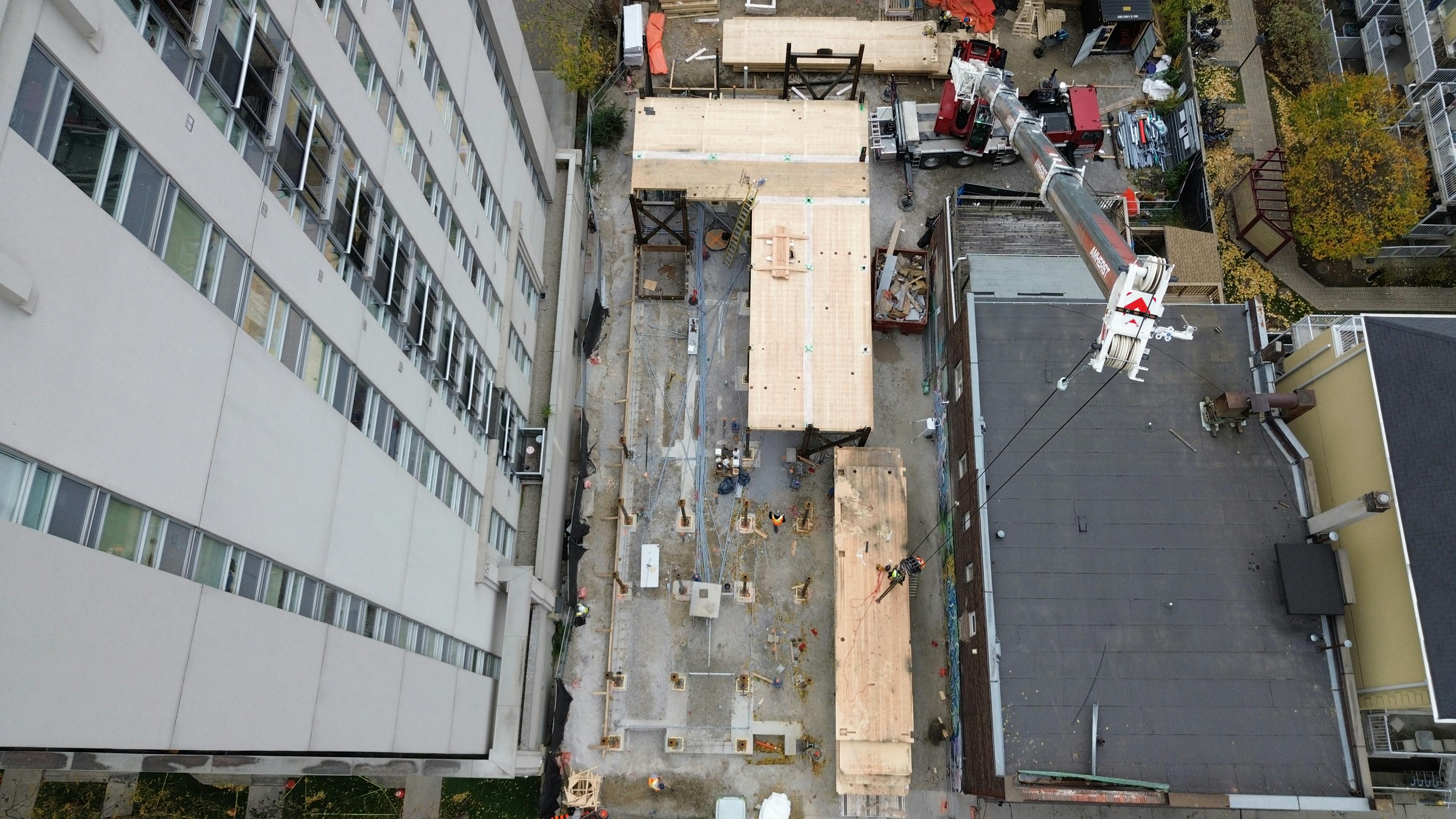 Top-down aerial view of 215 Wellesley St partially completed with mass timber platforms supported by steel beams. Workers and equipment are visible throughout the site. The construction is bordered by a tall residential building on the left and other buildings on the right.