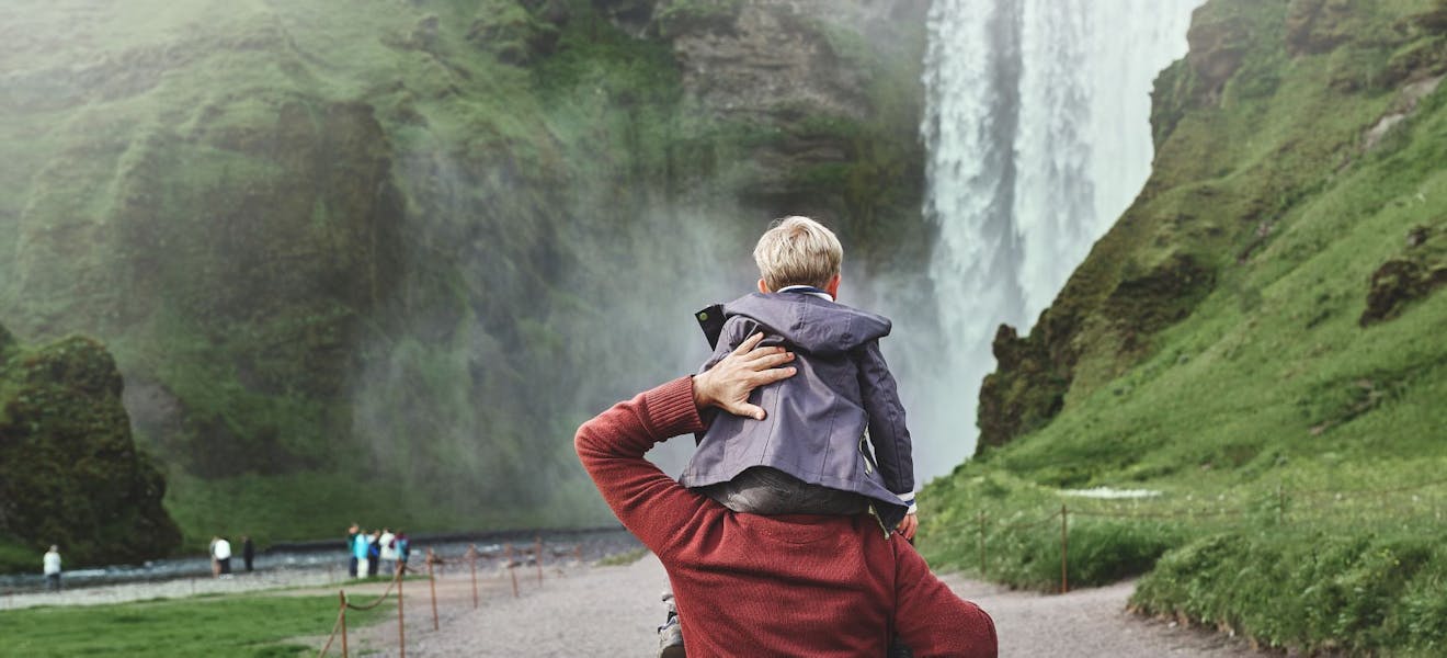 Family in vacation traveling in Iceland, father carries a small son on his shoulders to the famous waterfalls of Skogafoss