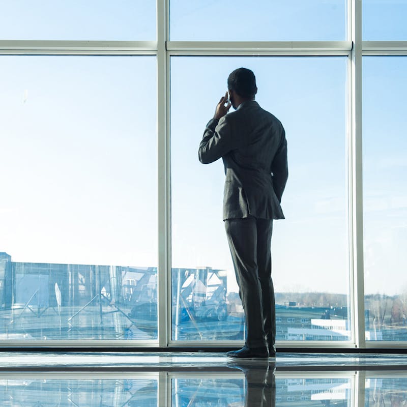 Maður í jakkafötum stendur við gólfsíða glugga og horfir út á meðan hann talar í farsíma