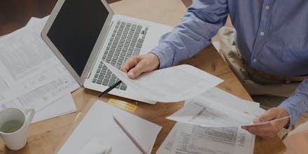Man in a blue shirt holding two tax sheets in front of a laptop