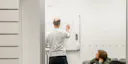 College-paitainen mies piirtää tussilla valkotaululle, toisen miehen tarkastellessa vieressä.