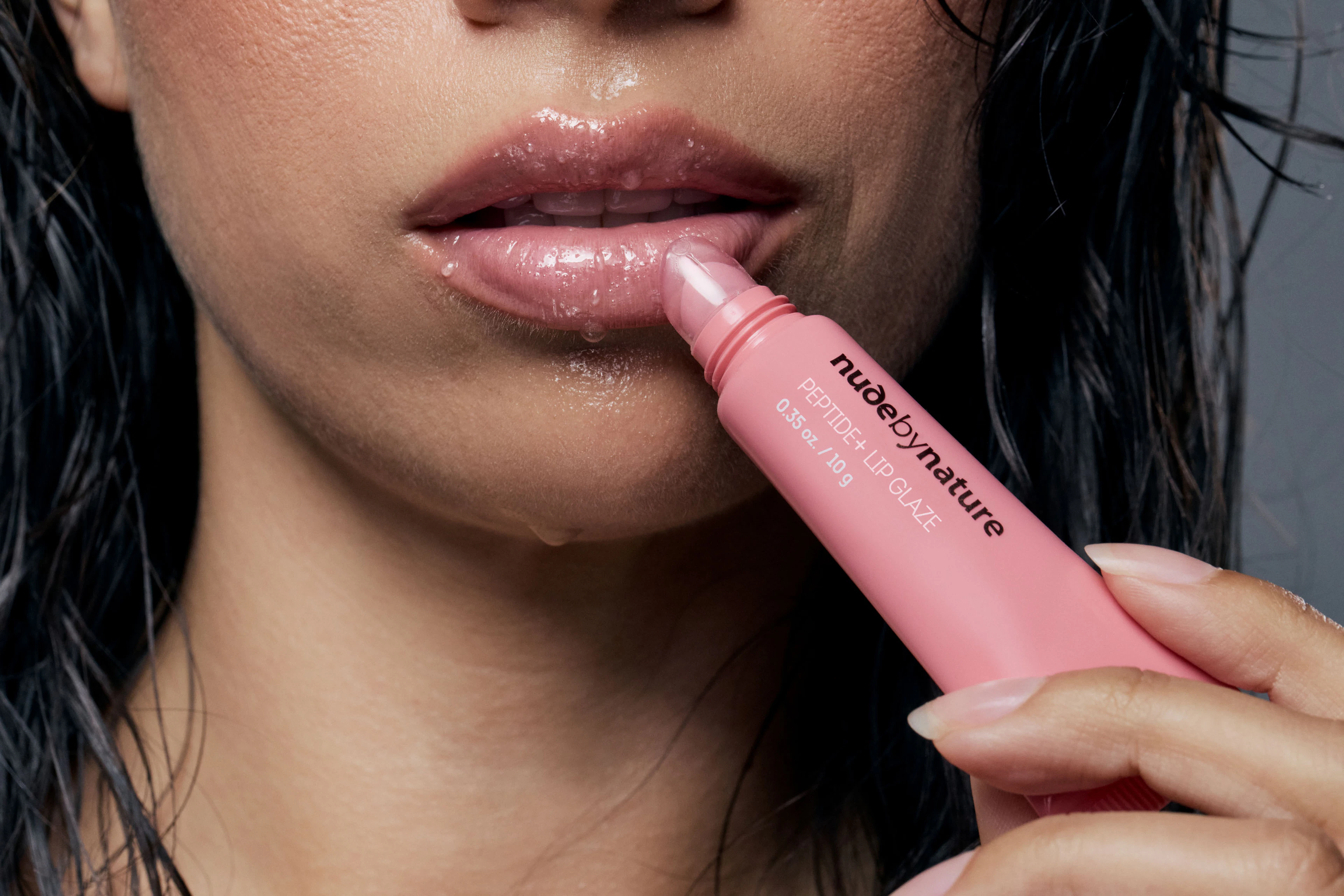 Extreme close up shot of woman applying glossy pink lip glaze to lips in studio for an editorial style Nude by Nature beauty photography campaign