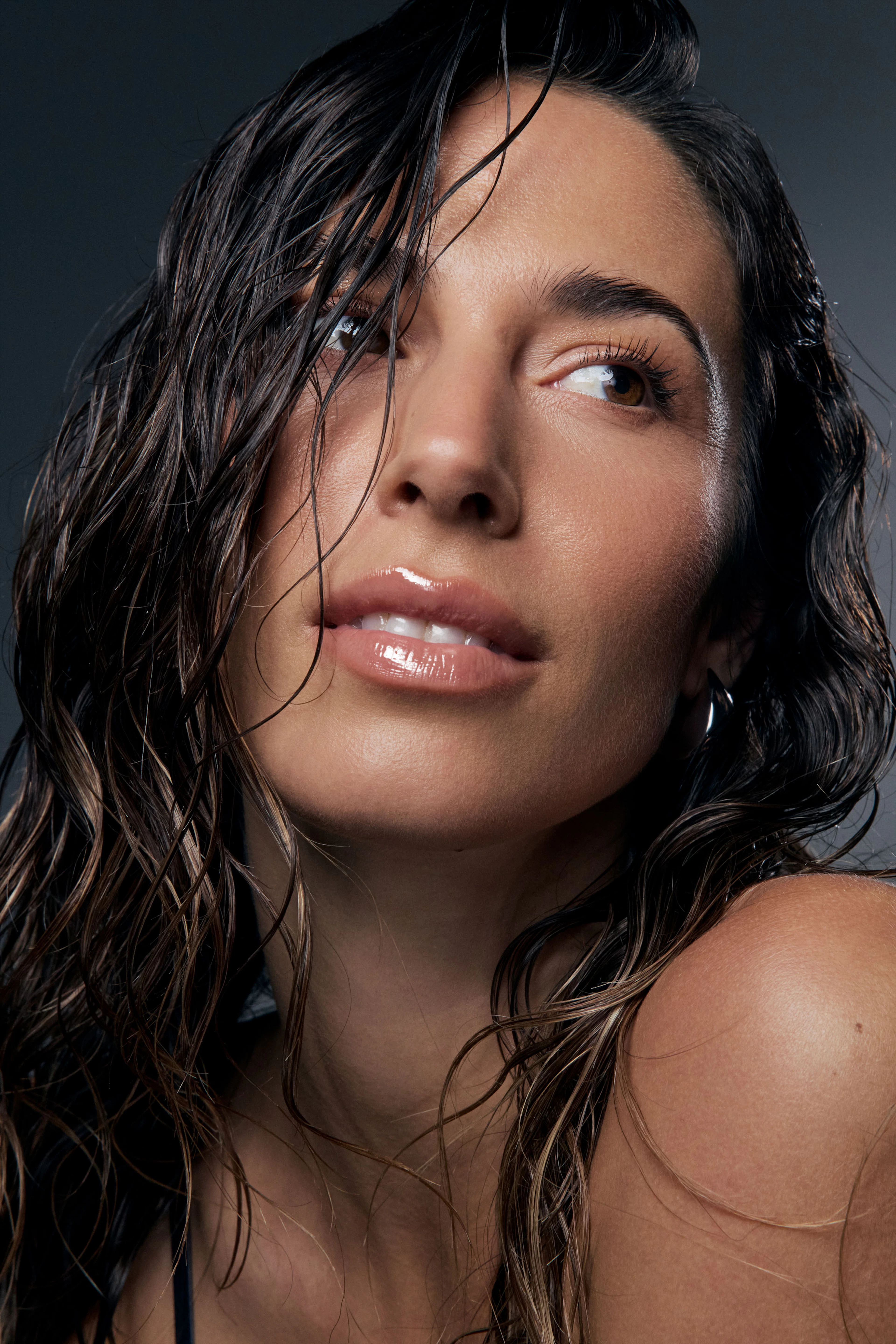 Woman with wet look hair wearing glossy pink lip glaze against a dark studio backdrop for a Nude by Nature beauty photography campaign