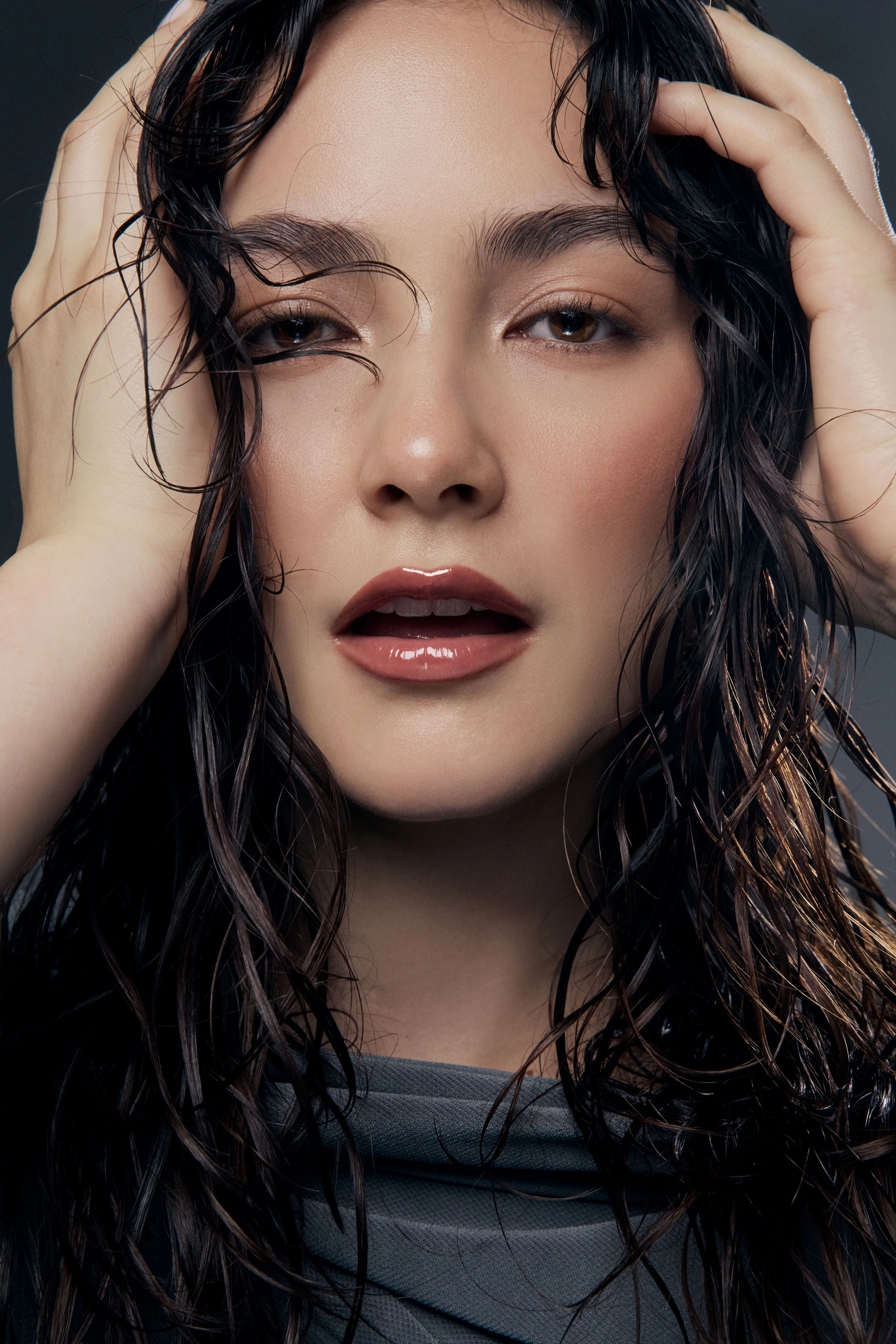 Woman with wet look hair wearing glossy pink lip glaze with hands on sides of head against dark studio backdrop for an editorial style Nude by Nature beauty photography campaign