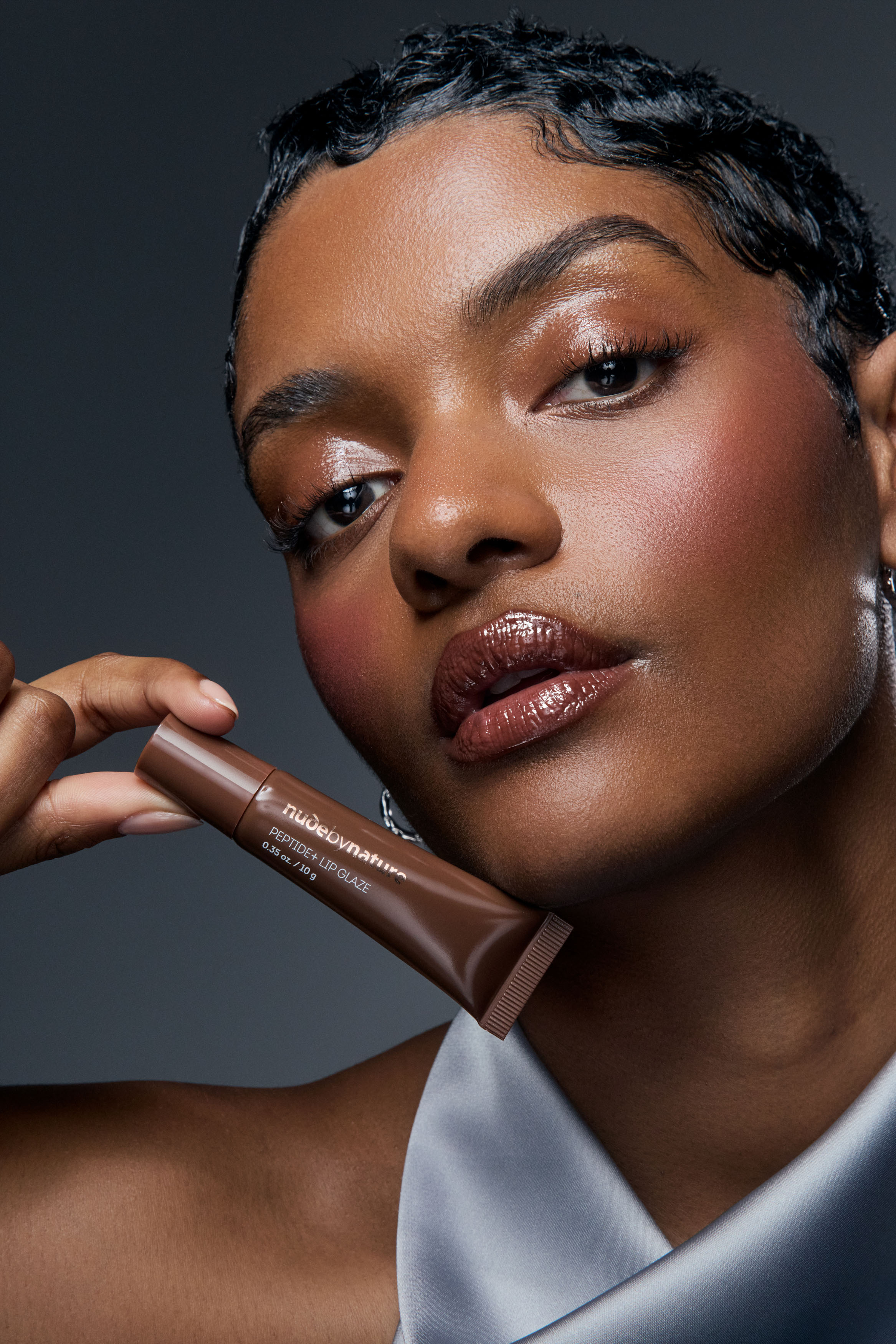 Creative beauty campaign photography for Nude by Nature Peptide+ Lip Glaze featuring a woman wearing glossy brown lip balm holding a product tube near her face in studio with moody lighting